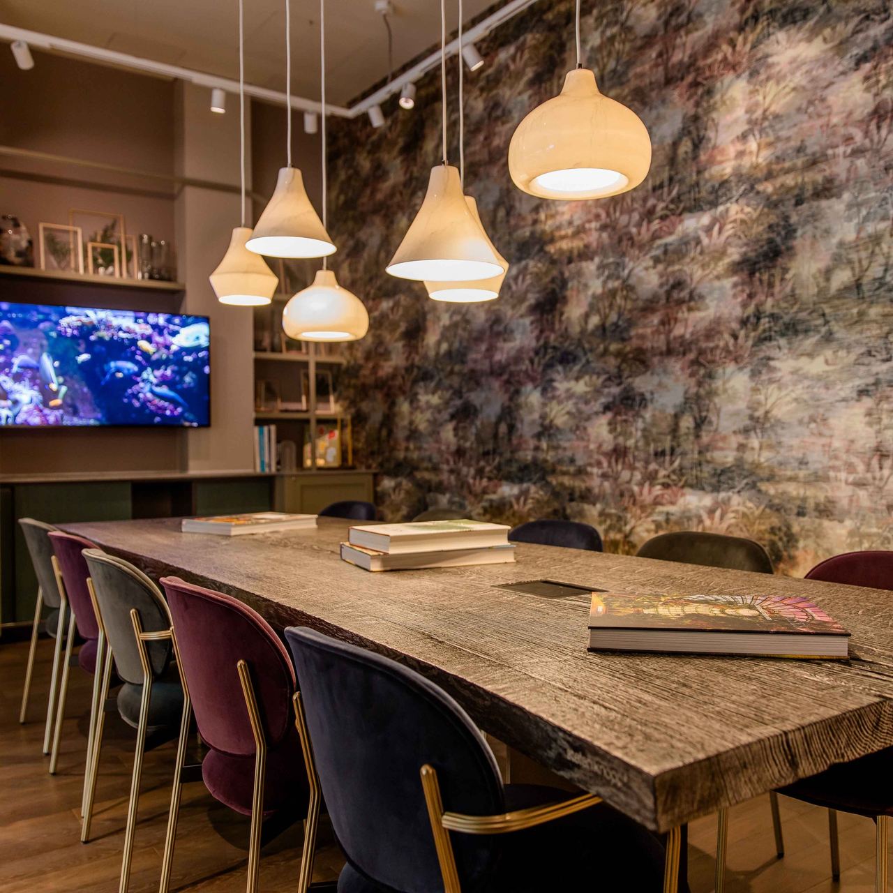 Motel One Konferenzraum mit langem Holztisch, mehreren farbigen Stühlen und warmen Hängelampen mit Kunst an der Wand und großem Bildschirm für Präsentationen.