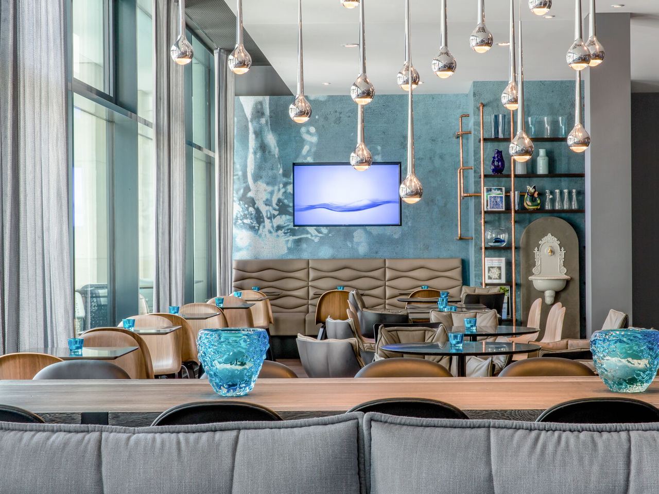 Motel One Lounge-Bereich mit gemütlichen Sitzgruppen, blauen Dekorelementen und großen Fenstern, die viel Tageslicht bieten. Stilvolle Pendelleuchten hängen von der Decke
