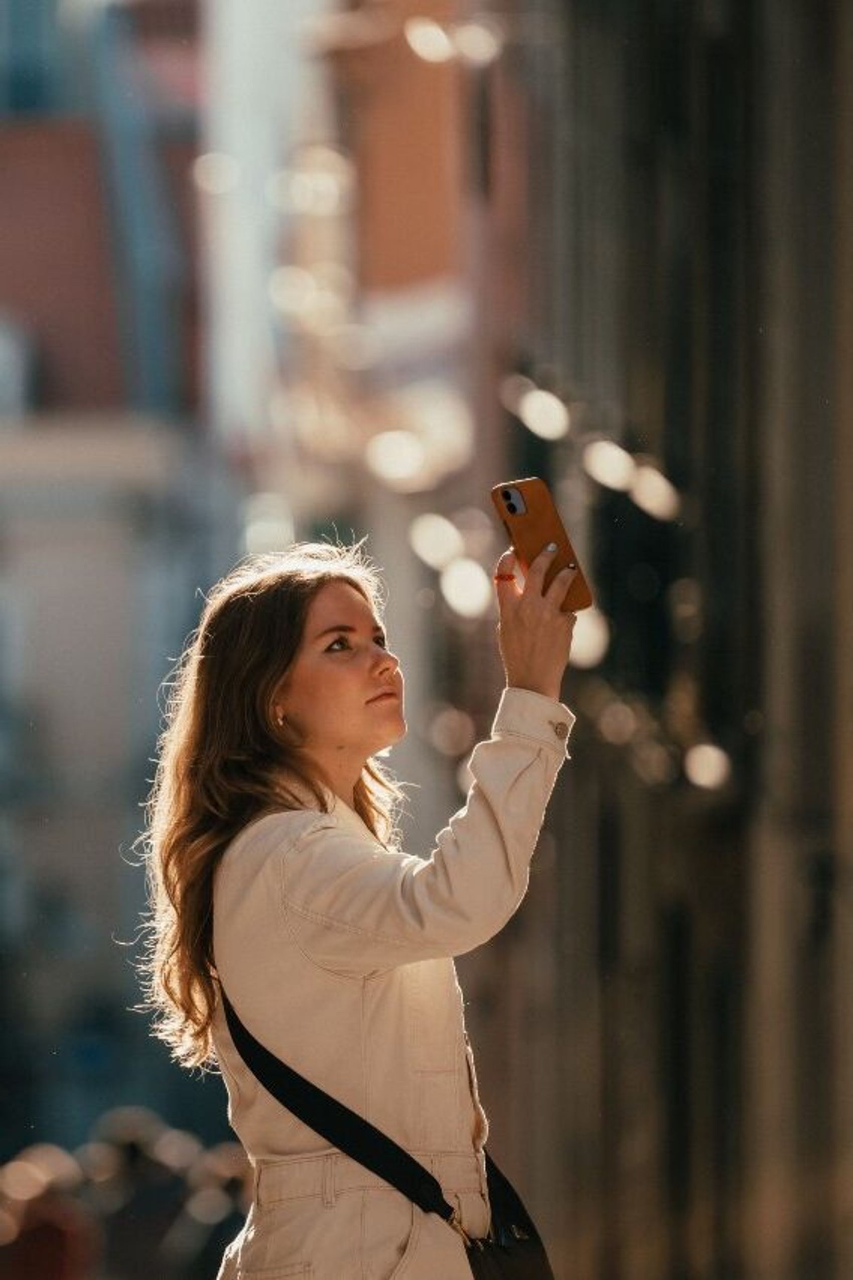 Junge Frau steht in einer sonnendurchfluteten Gasse und hält ihr Smartphone zum Fotografieren nach oben, wirkt konzentriert und neugierig.