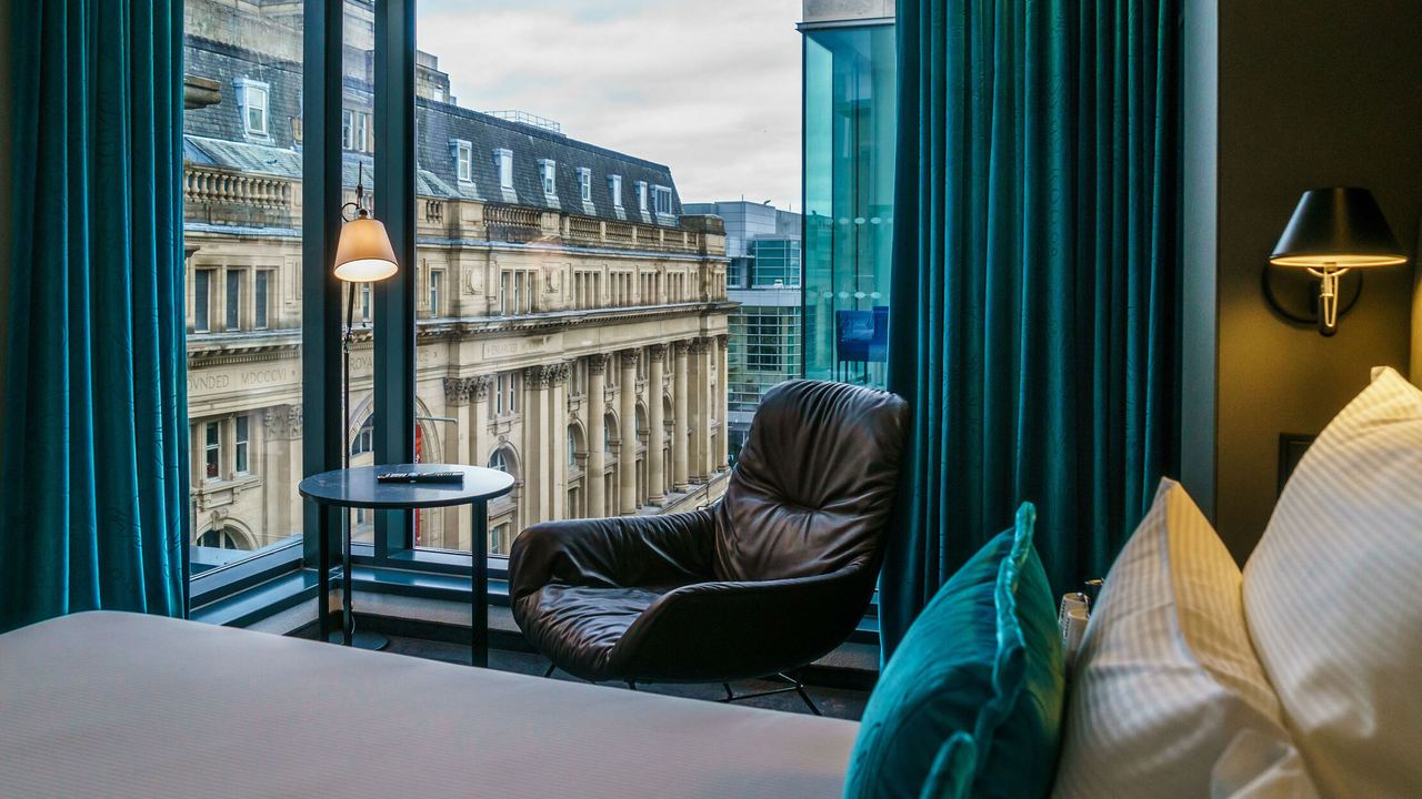 Blick aus einem Hotelzimmer mit gemütlichem Sessel und dunklen Vorhängen auf historische Stadtarchitektur, die durch große Panoramafenster sichtbar ist. Ruhige Atmosphäre für entspannte Momente mit urbanem Flair.