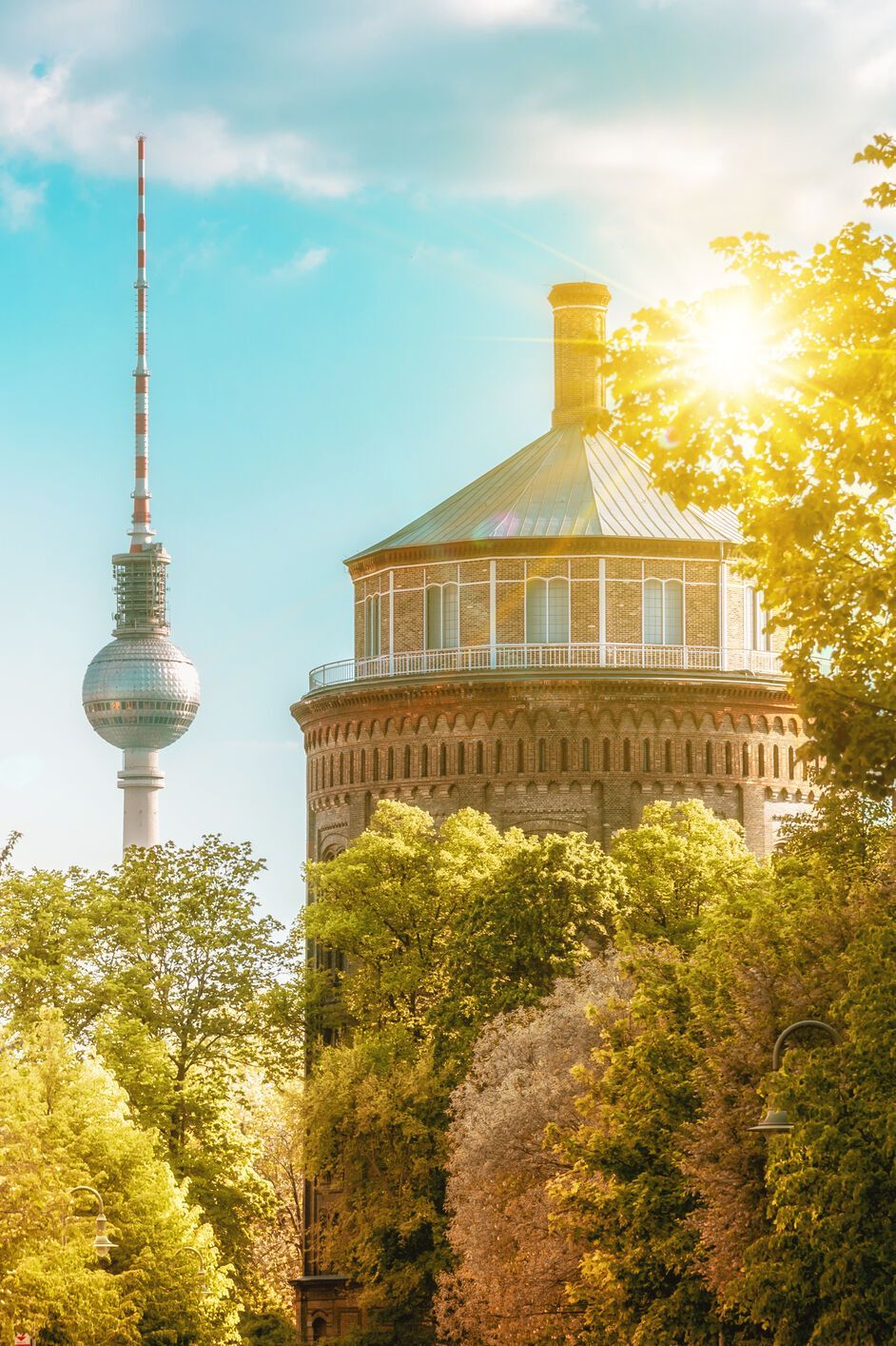 Sonnenbeschienene Straßenansicht in Berlin mit grünen Bäumen, historischen Gebäuden und dem Fernsehturm im Hintergrund, die eine einladende urbane Atmosphäre vermittelt.