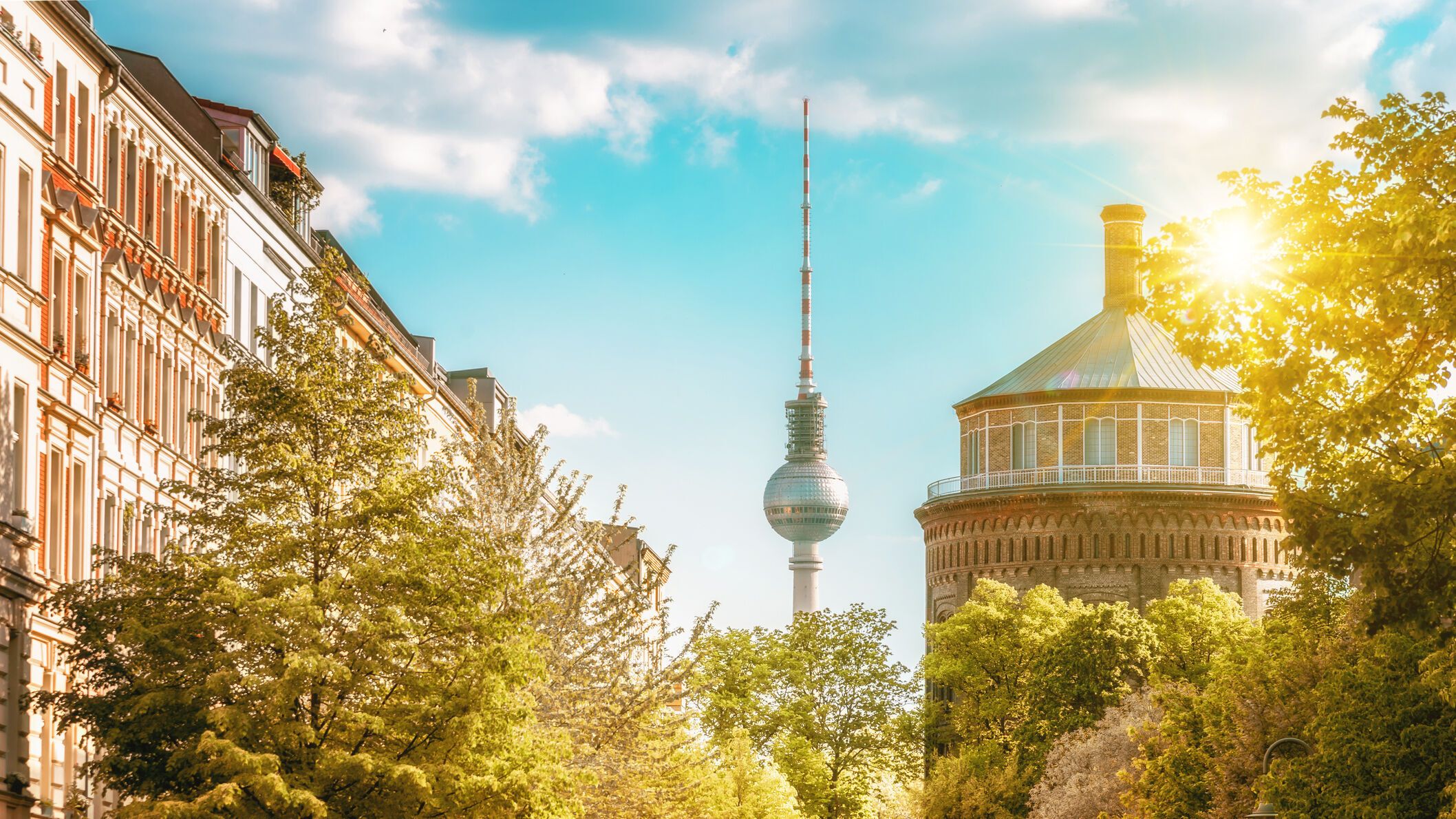 Sonnenbeschienene Straßenansicht in Berlin mit grünen Bäumen, historischen Gebäuden und dem Fernsehturm im Hintergrund, die eine einladende urbane Atmosphäre vermittelt.