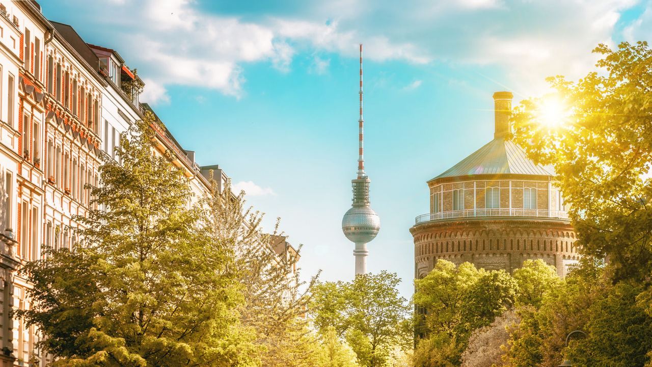 Sonnenbeschienene Straßenansicht in Berlin mit grünen Bäumen, historischen Gebäuden und dem Fernsehturm im Hintergrund, die eine einladende urbane Atmosphäre vermittelt.