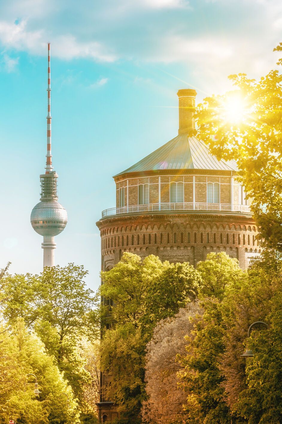 Sonnenbeschienene Straßenansicht in Berlin mit grünen Bäumen, historischen Gebäuden und dem Fernsehturm im Hintergrund, die eine einladende urbane Atmosphäre vermittelt.