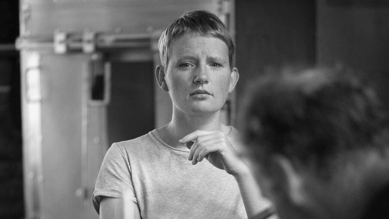 Junge Frau mit kurzem Haar schaut nachdenklich und konzentriert, im Vordergrund unscharfe Person, die ein Holzstück bearbeitet, Atmosphäre wirkt ruhig und introspektiv.