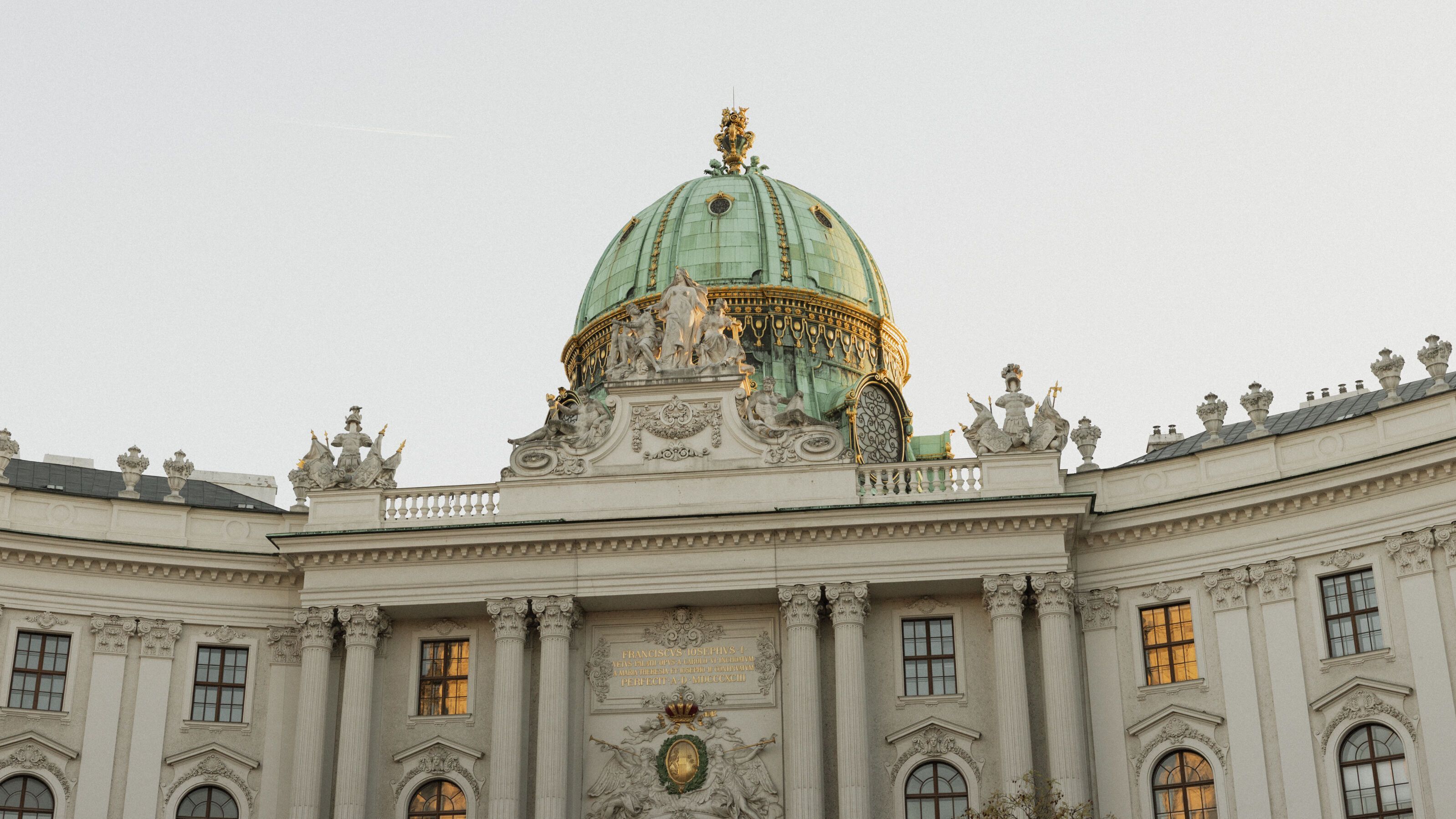 Historisches Gebäude mit einer großen grünen Kuppel, reich verzierten Fassaden und beleuchteten Fenstern, aufgenommen bei Tageslicht. Symbol für Kultur und Architektur in der Stadt.