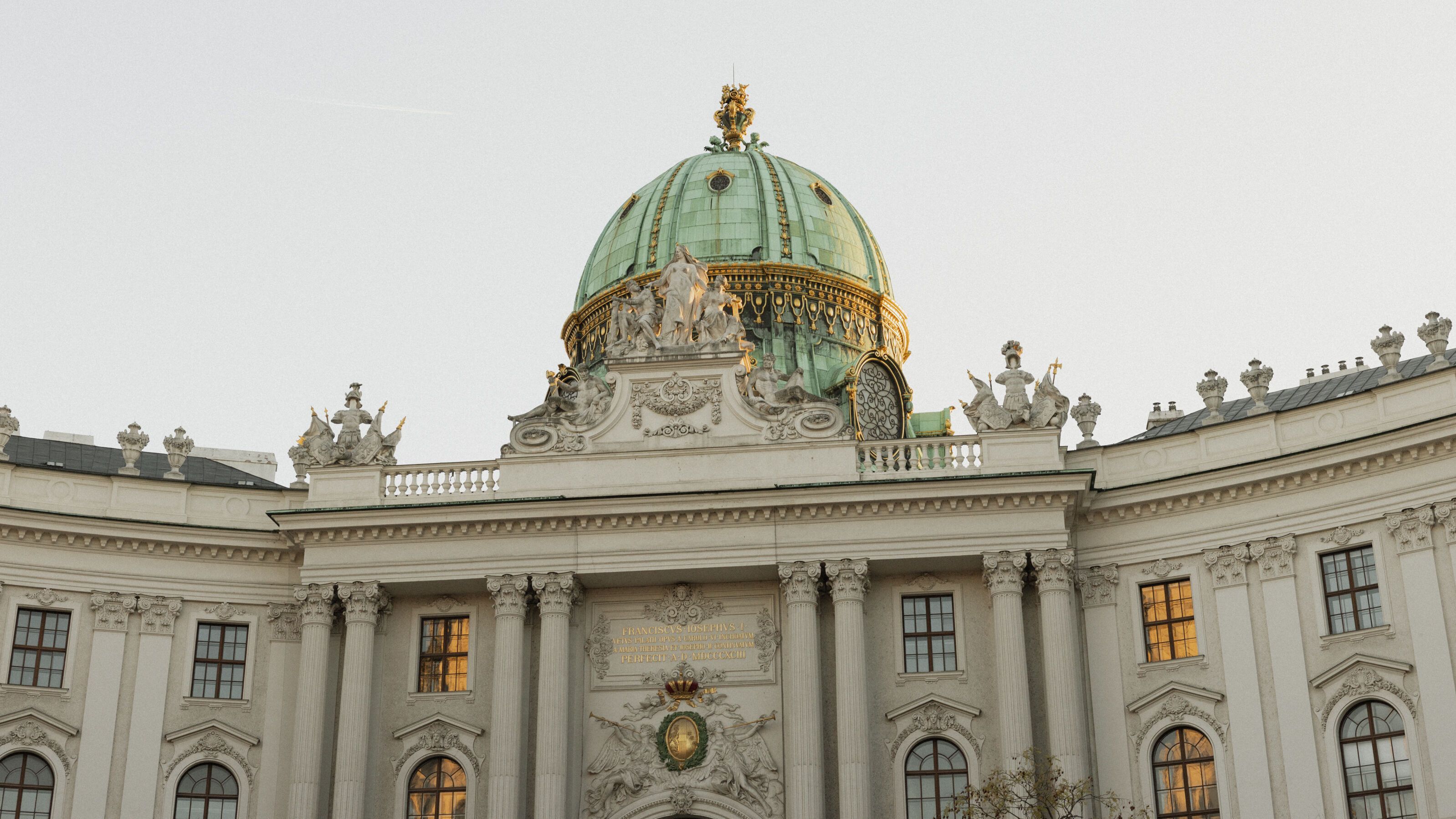 Historisches Gebäude mit einer großen grünen Kuppel, reich verzierten Fassaden und beleuchteten Fenstern, aufgenommen bei Tageslicht. Symbol für Kultur und Architektur in der Stadt.