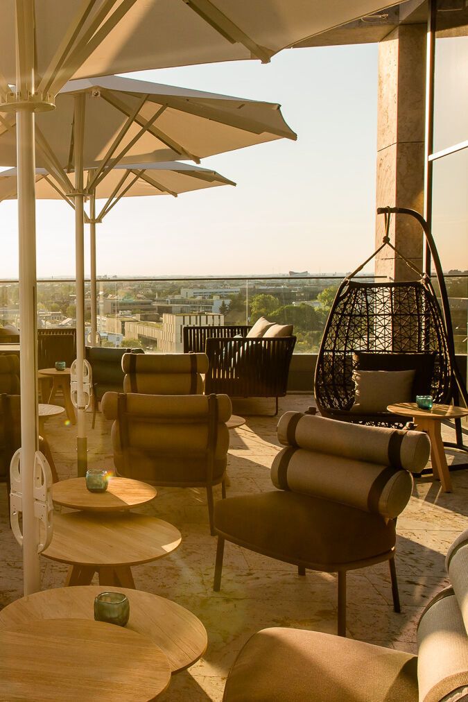 Sonnige Dachterrasse mit bequemen Sitzmöbeln, Tischen und großen Sonnenschirmen, Blick auf eine Stadtlandschaft im Hintergrund.