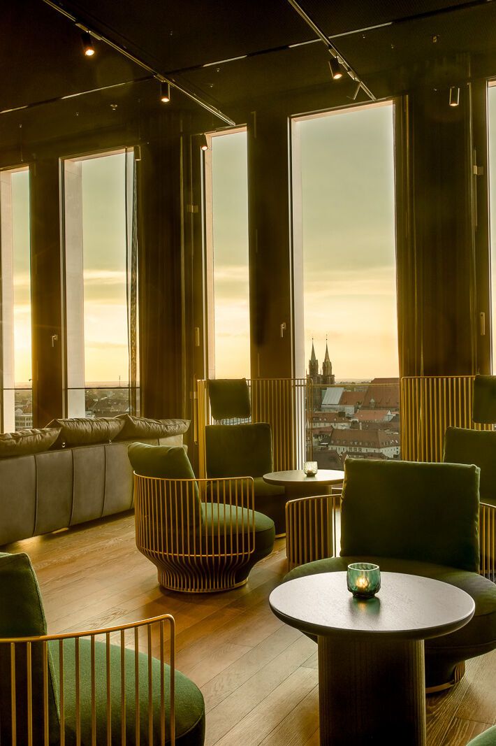 One Lounge mit bequemen Sesseln, großen Panoramafenstern und Sonnenuntergangsblick über eine Stadt mit historischen Türmen