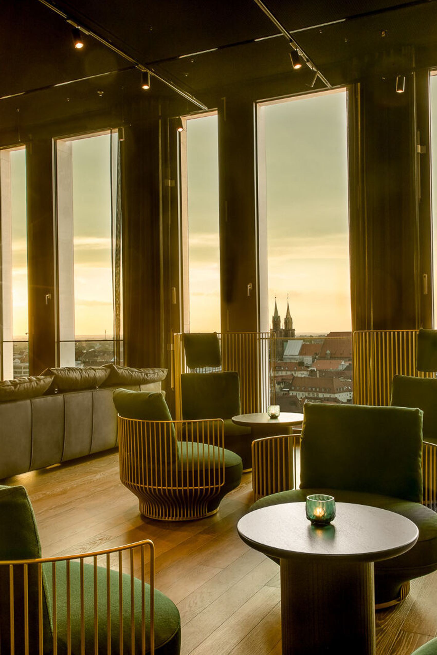 One Lounge mit bequemen Sesseln, großen Panoramafenstern und Sonnenuntergangsblick über eine Stadt mit historischen Türmen
