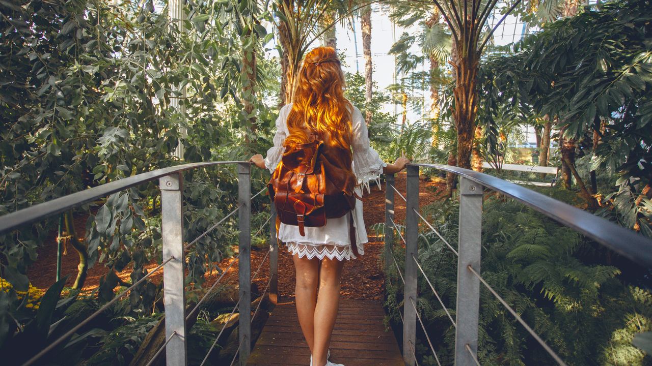 Frau mit Rucksack geht auf einer Holzbrücke durch einen üppigen, sonnendurchfluteten tropischen Botanischen Garten.