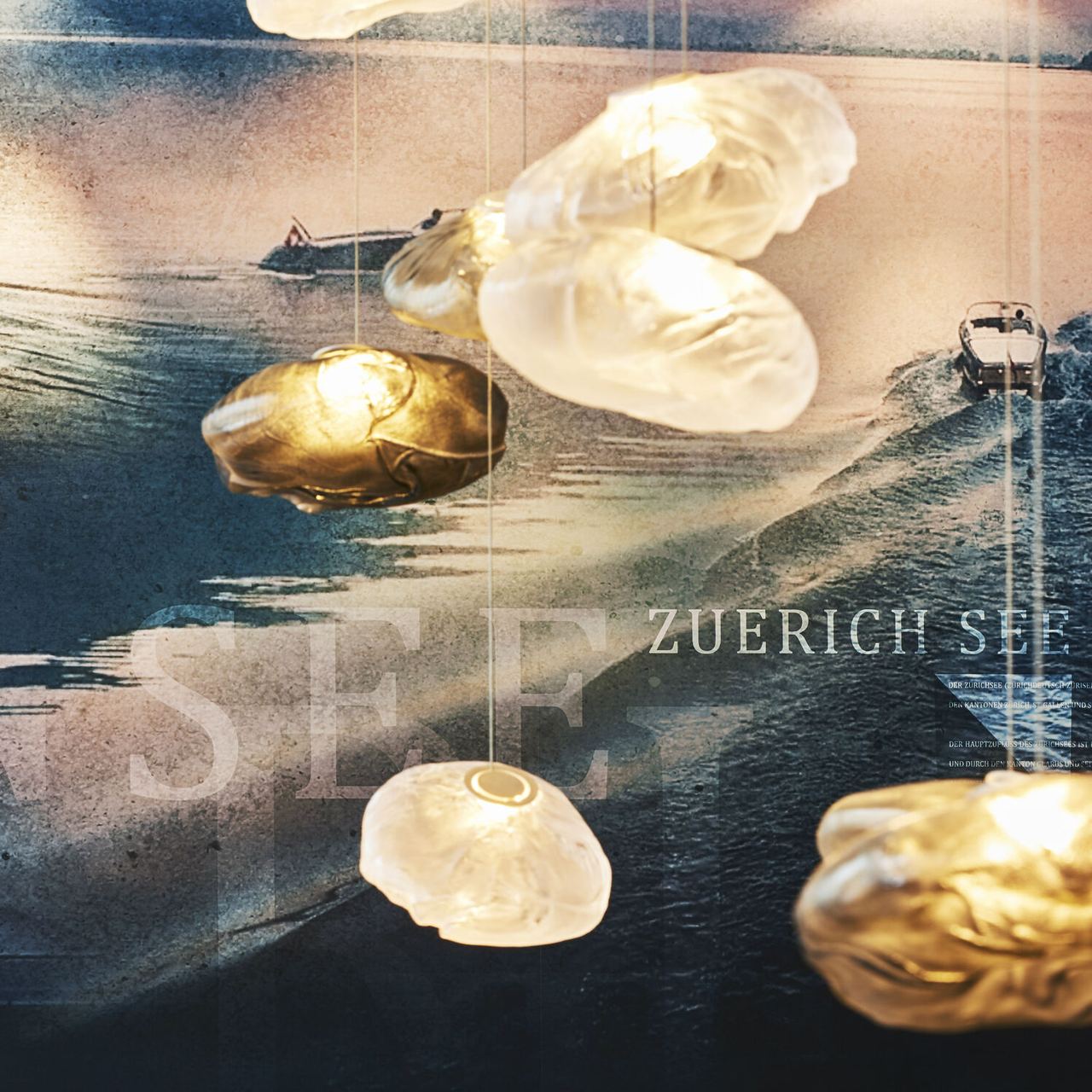 Beleuchtete, organisch geformte Lampen schweben vor einem stimmungsvollen Bild des Zürichsees bei Sonnenuntergang mit Booten auf dem Wasser.