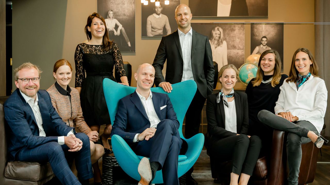 Acht beruflich gekleidete Personen sitzen und stehen in einem einladenden Büroambiente und zeigen Selbstbewusstsein und Teamzusammenhalt.
