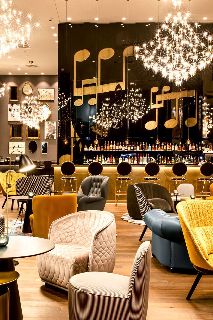 Motel One Hotelbar mit modernem, gemütlichem Interieur, farbenfrohen Sesseln und Sofas, stilvollen Tischen sowie musikalischer Wanddekoration.