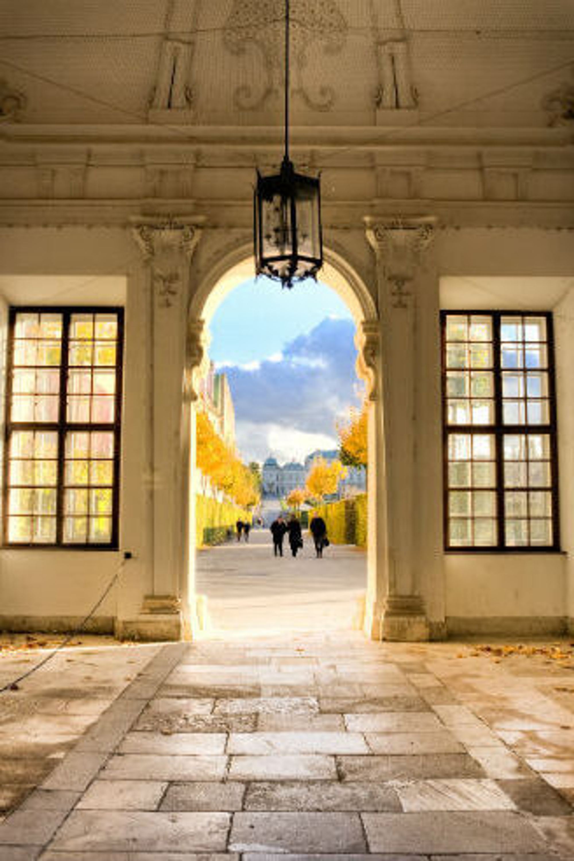 Blick durch einen prunkvollen, mit Statuen geschmückten Arkadengang auf einen herbstlichen Parkweg mit Menschen, der zu einem historischen Schloss im Hintergrund führt. Helle, warme Lichtstimmung und klarer Himmel schaffen eine einladende Atmosphäre.