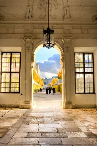 Blick durch einen prunkvollen, mit Statuen geschmückten Arkadengang auf einen herbstlichen Parkweg mit Menschen, der zu einem historischen Schloss im Hintergrund führt. Helle, warme Lichtstimmung und klarer Himmel schaffen eine einladende Atmosphäre. Blick durch einen prunkvollen, mit Statuen geschmückten Arkadengang auf einen herbstlichen Parkweg mit Menschen, der zu einem historischen Schloss im Hintergrund führt. Helle, warme Lichtstimmung und klarer Himmel schaffen eine einladende Atmosphäre.