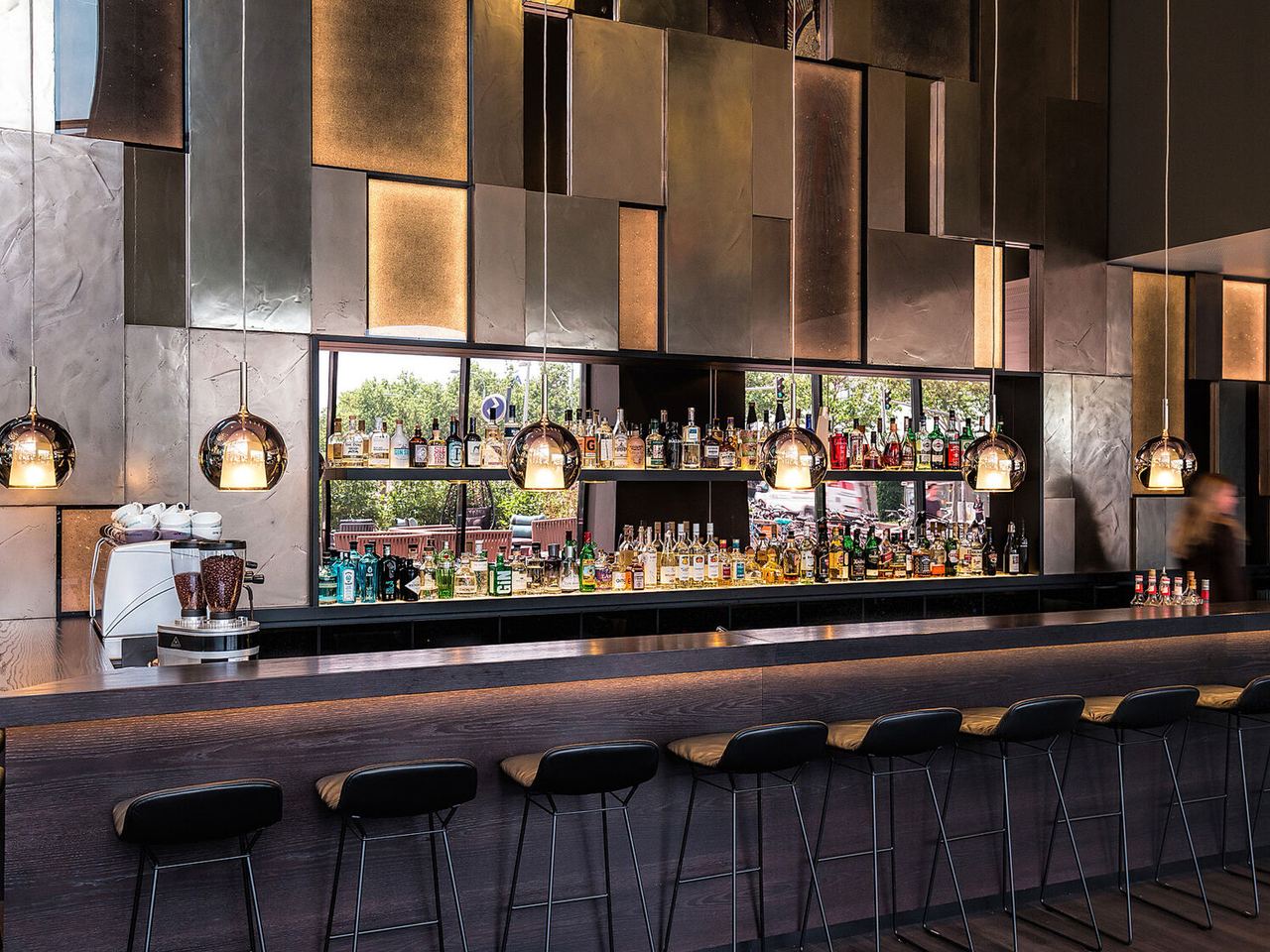 Motel One Hotelbar mit langem, dunklem Tresen und sechs schwarzen Barhockern, beleuchtet von modernen Hängeleuchten. Im Hintergrund sind vielfältige Spirituosen und große Fenster mit Außenblick zu sehen.
