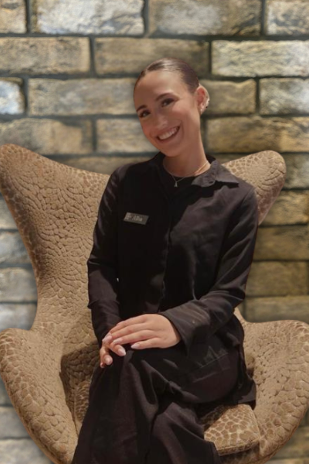 Junge Hotelmitarbeiterin in schwarzer Uniform sitzt lächelnd auf einem bequemen Sessel vor einer Steinwand, wirkt freundlich und einladend.