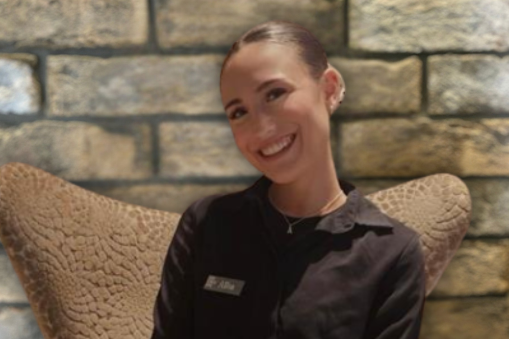 Junge Hotelmitarbeiterin in schwarzer Uniform sitzt lächelnd auf einem bequemen Sessel vor einer Steinwand, wirkt freundlich und einladend. Junge Hotelmitarbeiterin in schwarzer Uniform sitzt lächelnd auf einem bequemen Sessel vor einer Steinwand, wirkt freundlich und einladend.