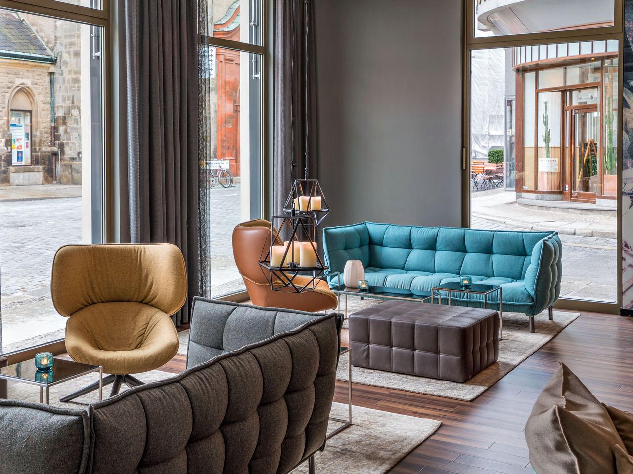 Motel One Lounge-Bereich mit gemütlichen Sitzmöbeln in gedeckten Farben, großen Fenstern mit Blick auf den ruhigen Außenbereich und warmem Holzfußboden.