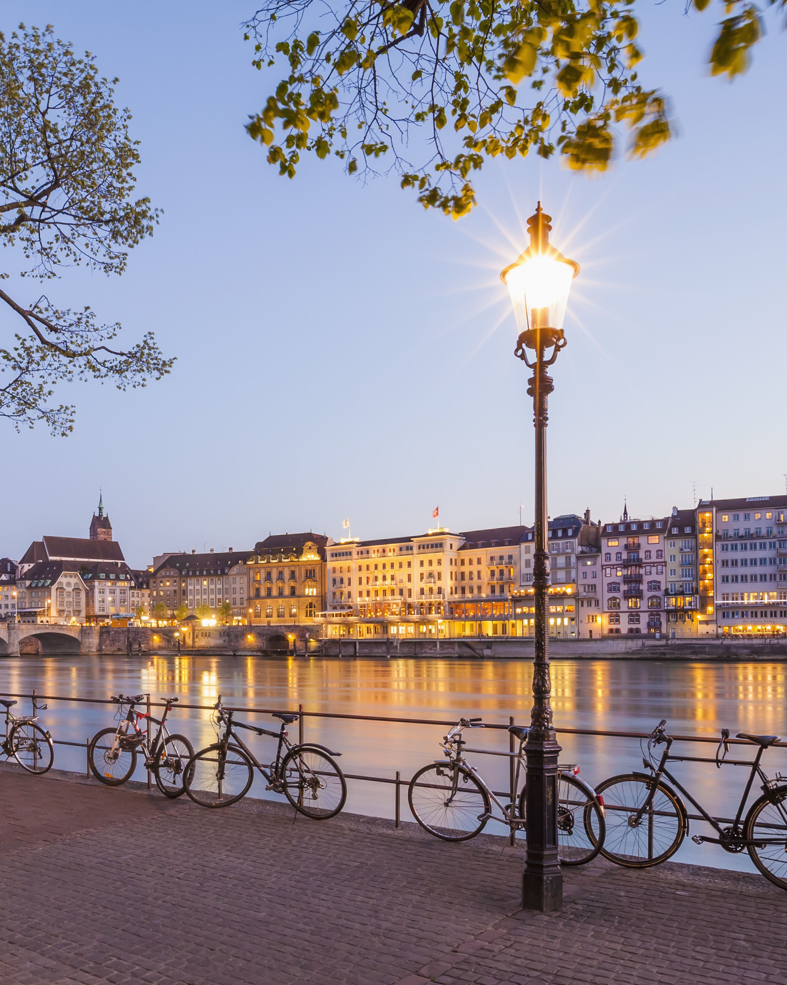 Uferpromenade am Fluss mit Fahrradständer und daran geparkten Fahrrädern, beleuchtete Stadt mit historischen Gebäuden und Brücke im Hintergrund bei Dämmerung.