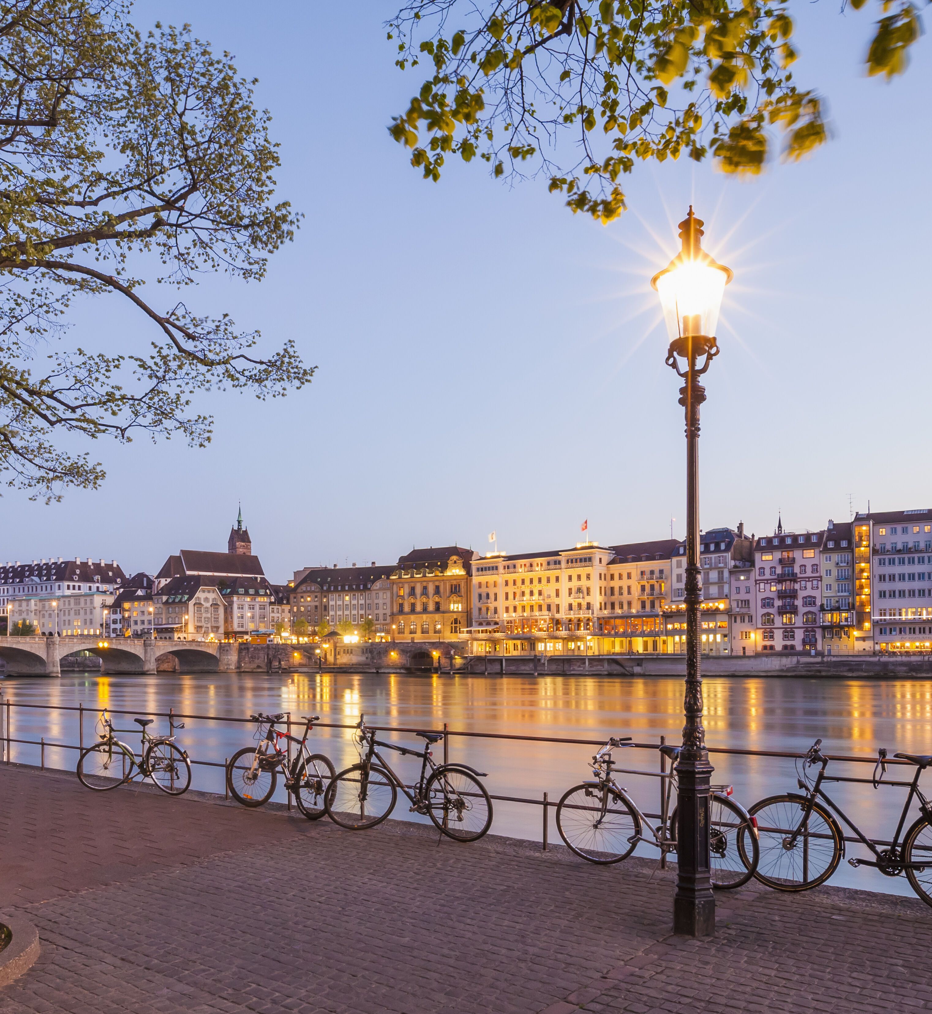Uferpromenade am Fluss mit Fahrradständer und daran geparkten Fahrrädern, beleuchtete Stadt mit historischen Gebäuden und Brücke im Hintergrund bei Dämmerung.
