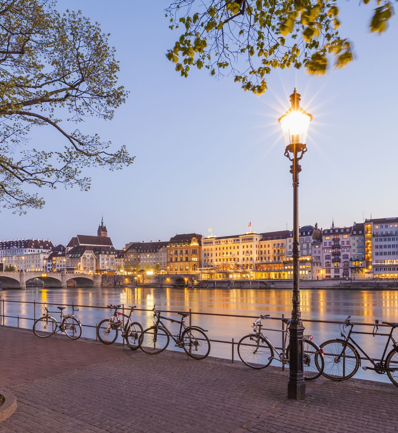 Uferpromenade am Fluss mit Fahrradständer und daran geparkten Fahrrädern, beleuchtete Stadt mit historischen Gebäuden und Brücke im Hintergrund bei Dämmerung.