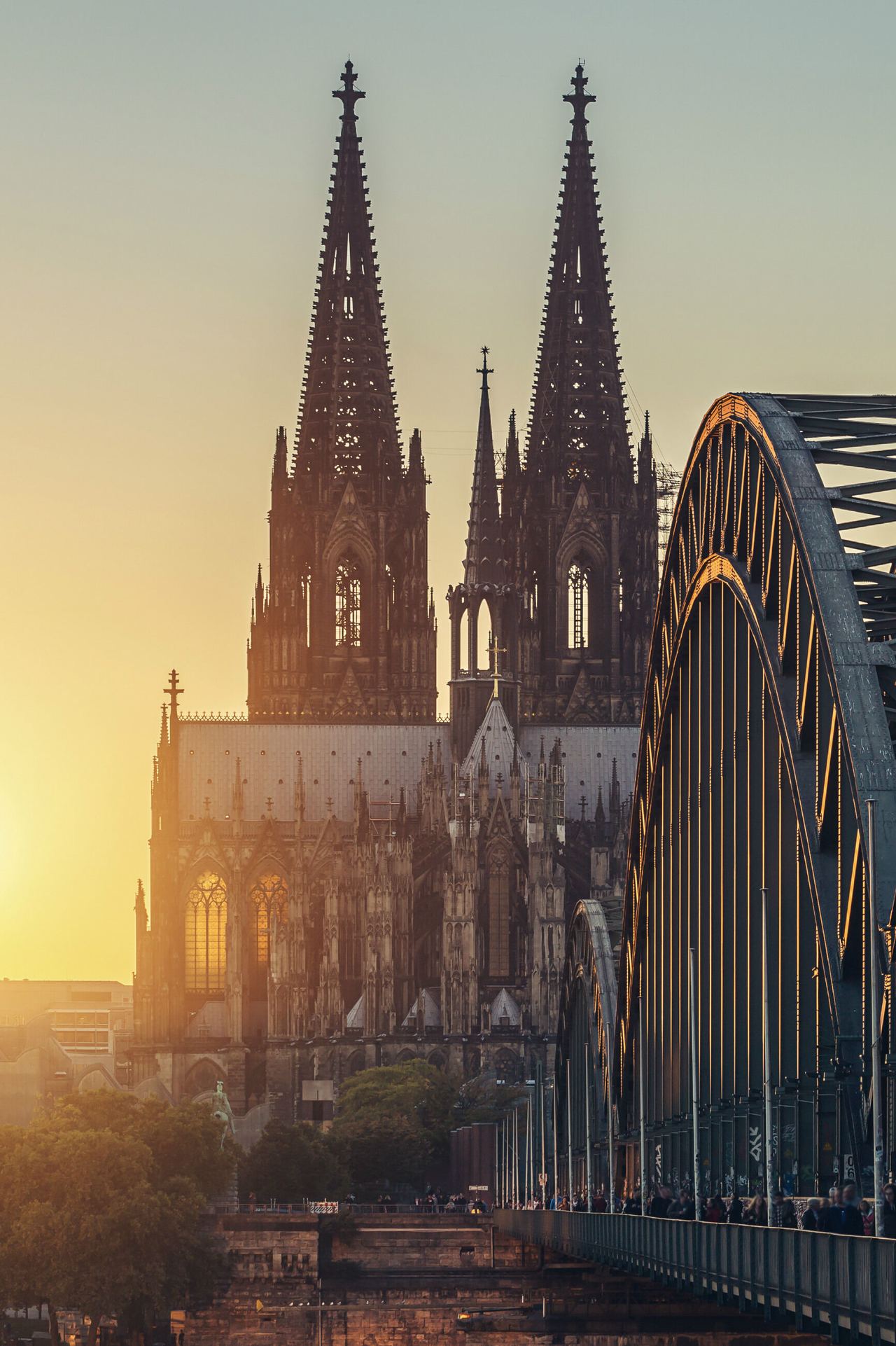 Blick auf den Kölner Dom bei Sonnenuntergang mit der markanten Eisenbahnbrücke im Vordergrund, auf der Menschen zu Fuß unterwegs sind.