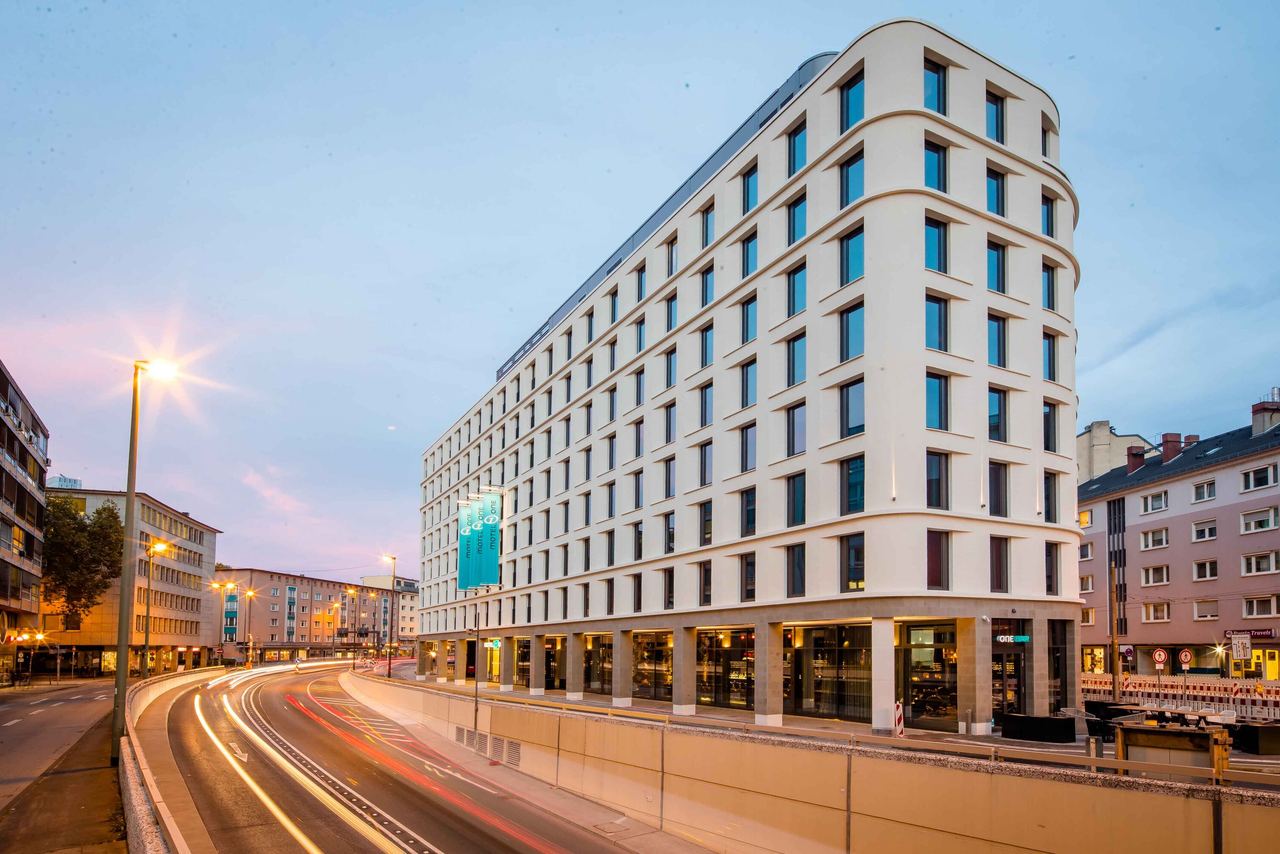 Motel One Hotelgebäude mit klaren Linien und großen Fenstern, gelegen an einer belebten Stadtstraße bei Abenddämmerung, mit Lichtspuren vorbeifahrender Fahrzeuge.