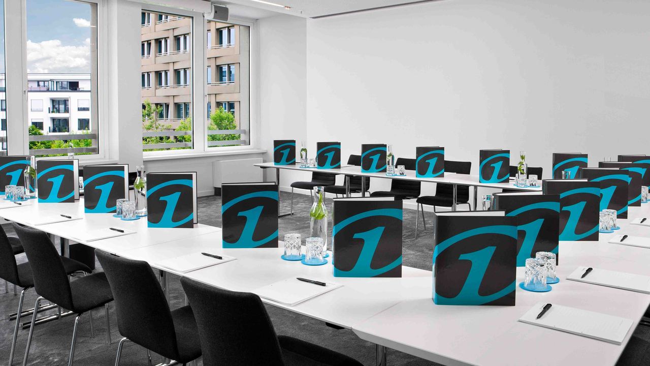 Motel One Konferenzraum mit großen Fenstern, Mobiliar und vorbereitetem Arbeitsplatz für mehrere Teilnehmer, ausgestattet mit Informationsmappen, Schreibblöcken und Wasserflaschen.
