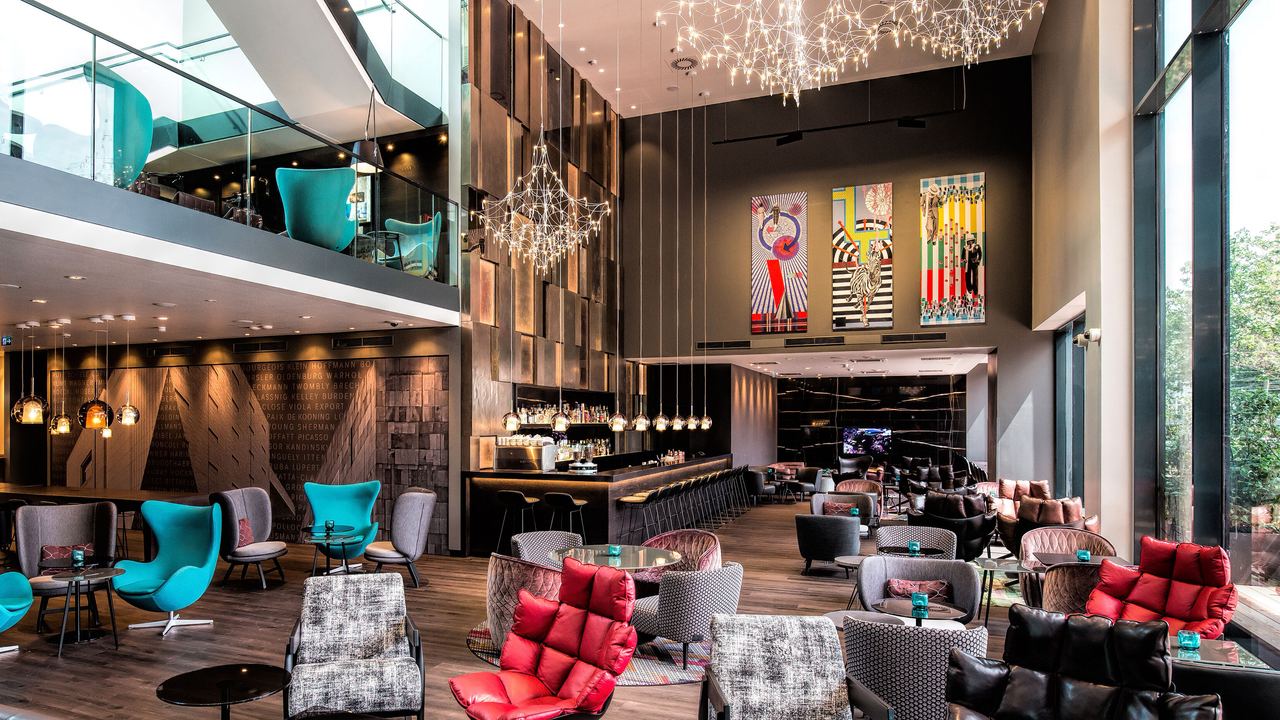 Motel One Hotel-Loungebereich mit hohen Decken, stilvollen Sesseln in verschiedenen Farben, großen Fensterfronten für viel Tageslicht und kunstvollen Deckenleuchten.