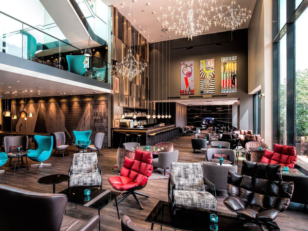 Motel One Hotel-Loungebereich mit hohen Decken, stilvollen Sesseln in verschiedenen Farben, großen Fensterfronten für viel Tageslicht und kunstvollen Deckenleuchten.