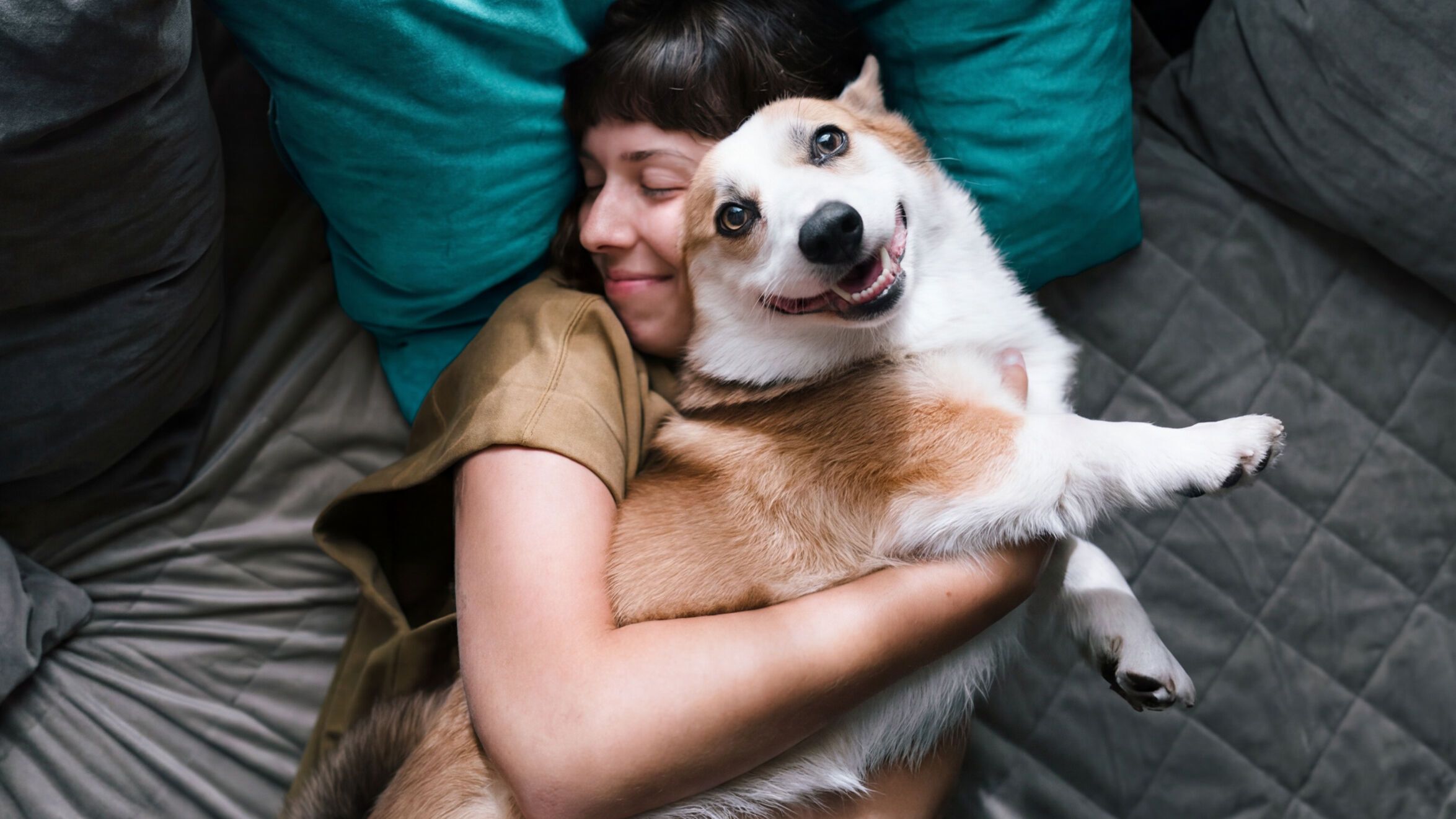 Frau liegt entspannt auf einem Sofa und umarmt glücklich ihren fröhlichen Hund, der in die Kamera blickt.