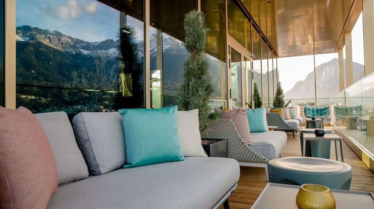 Balkon mit bequemen Sitzmöbeln und bunten Kissen, umgeben von Glaswänden, mit spektakulärem Blick auf alpine Berglandschaft bei Tageslicht.