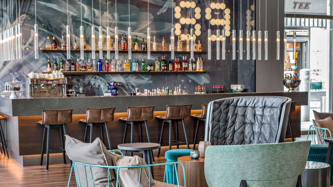 Motel One Hotelbar mit stilvollen Sitzgelegenheiten, gemütlichen Sesseln und Barhockern. Dezente Beleuchtung und ein gut bestücktes Getränkeangebot.