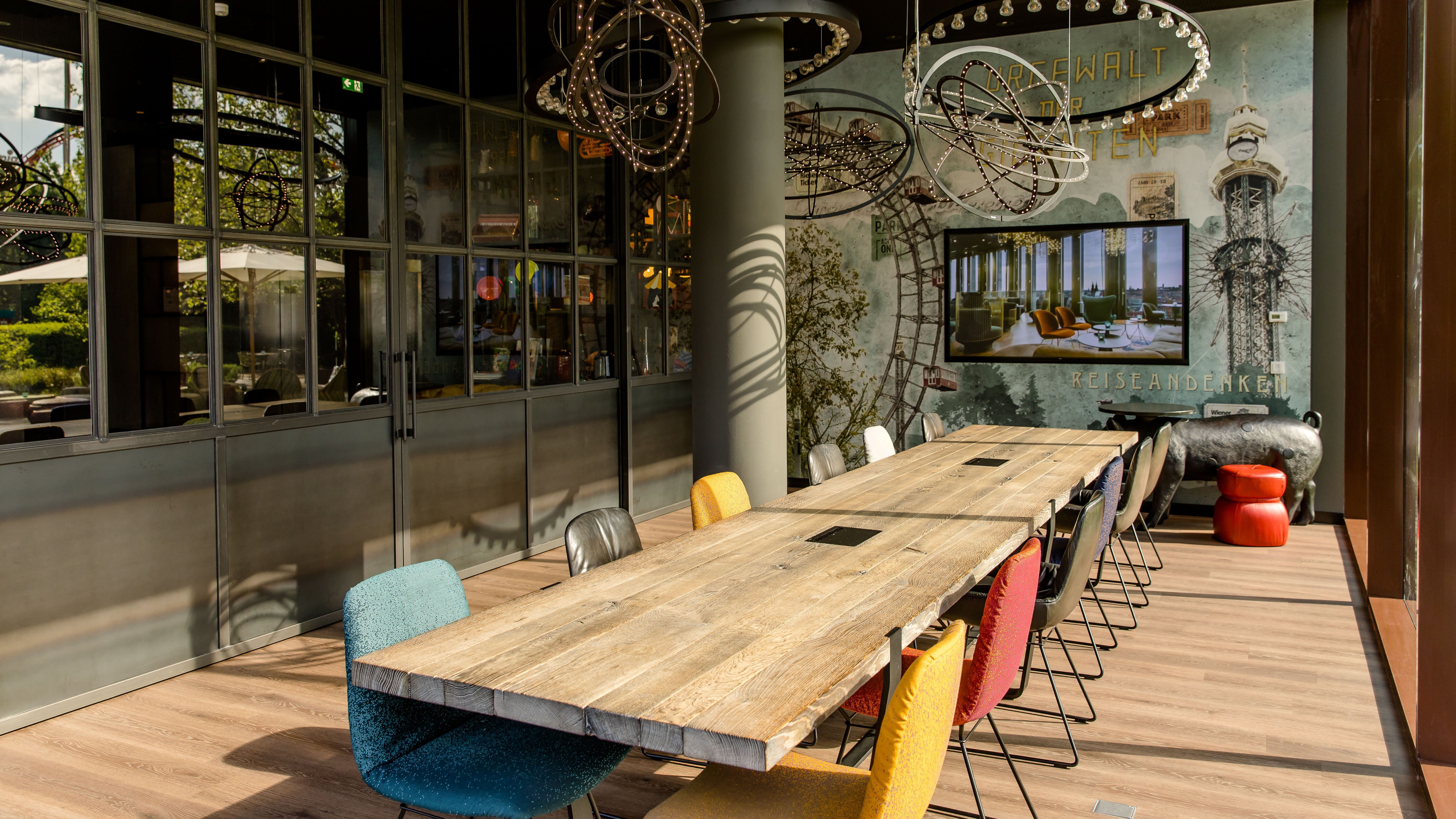 Motel One Besprechungsraum mit langem Holztisch, bunten Stühlen und großen Fenstern, die viel Tageslicht hereinlassen. An der Wand Kunstobjekte und ein Bildschirm für Präsentationen. 