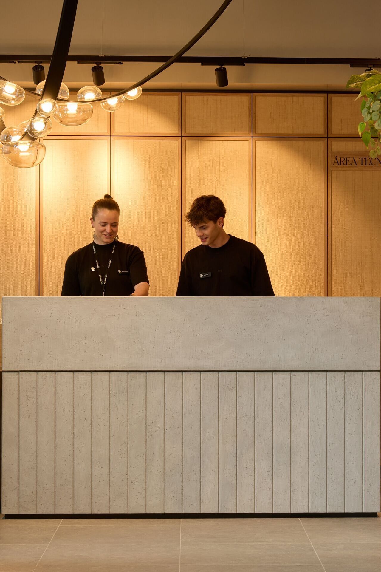 Empfangsbereich im The Cloud One Hotel mit zwei freundlichen Mitarbeitenden am modernen Tresen, warmem Licht und Pflanzen, die eine einladende Atmosphäre schaffen.