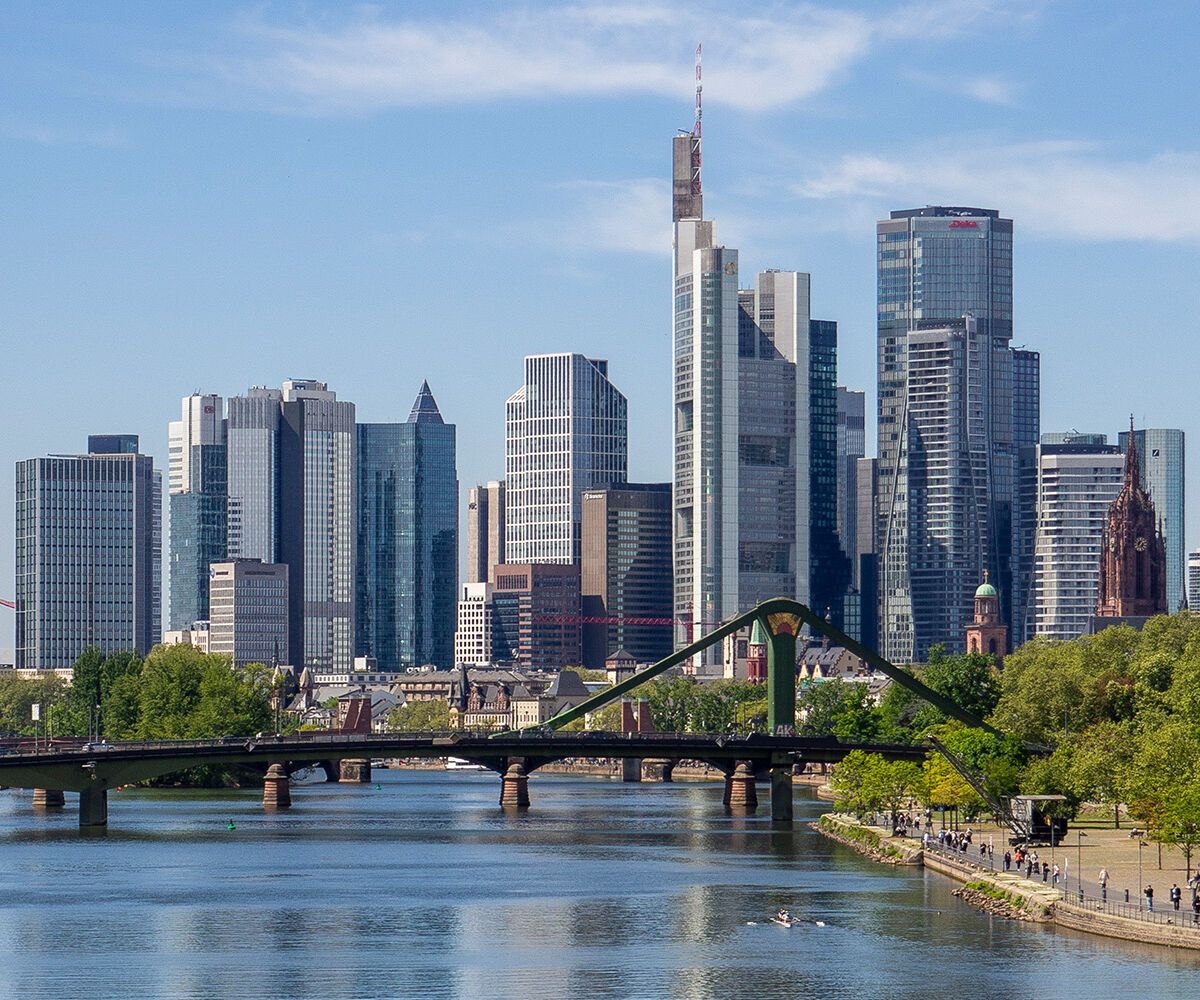 Panorama der Frankfurter Skyline mit modernen Wolkenkratzern, Fluss im Vordergrund und grünen Uferbereichen bei klarem Himmel.