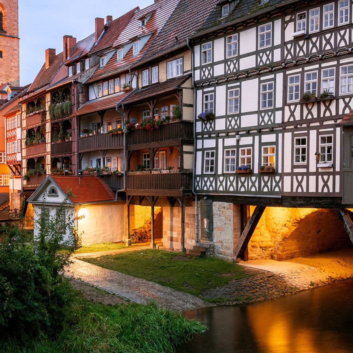 Alt-Text: Historische Fachwerkhäuser entlang eines Flusses mit beleuchteten Bögen und im Hintergrund ein hoher Kirchturm im warmen Abendlicht.