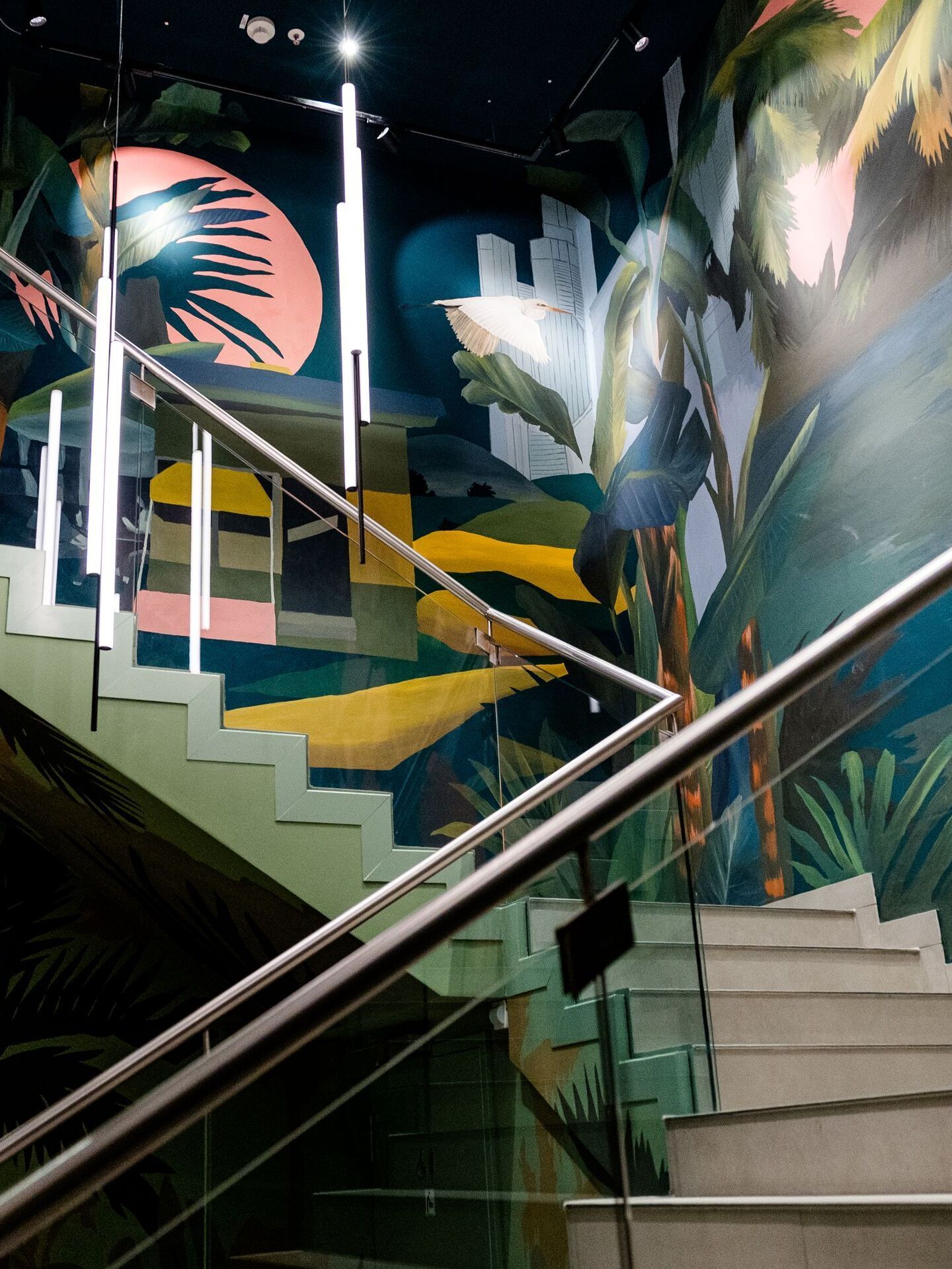 Bunte Wendeltreppe mit tropischen Wandgemälden in lebendigen Farben, die eine entspannte, exotische Atmosphäre schaffen. Edelstahl-Geländer reflektieren das Licht, das die Szenen sanft beleuchtet.