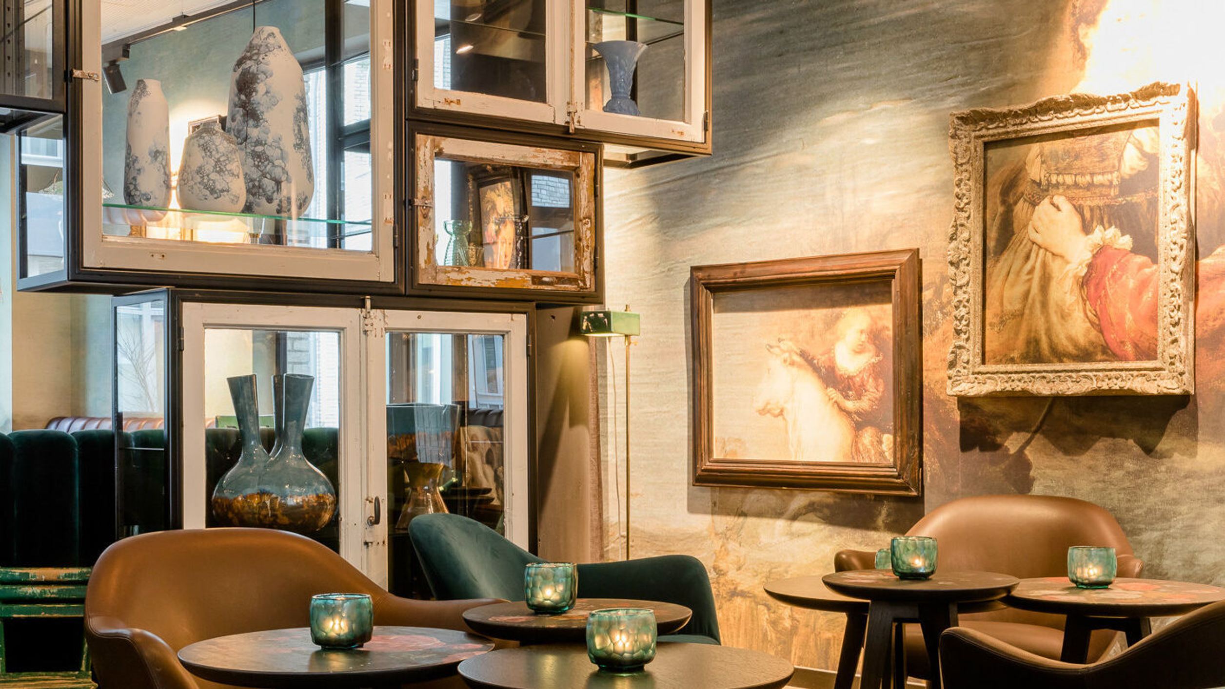 Motel One Lounge mit Leder- und Samtsesseln, runden Tischen und stimmungsvoller Kerzenbeleuchtung. Wände dekoriert mit klassischen Gemälden, Regale mit Vasen und dekorativen Objekten aus Upcycling Material.