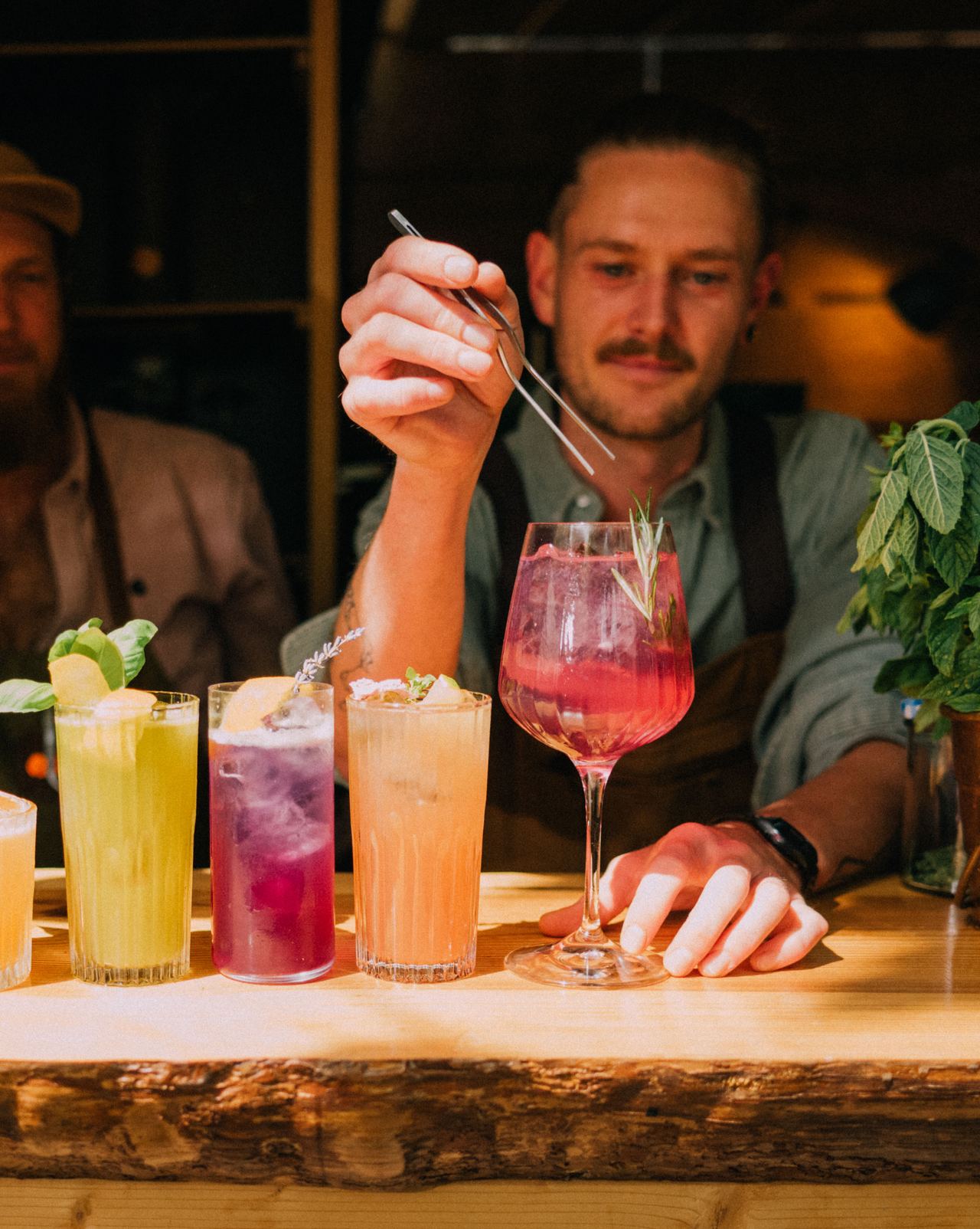 Zwei Barkeeper bereiten an einer Bar fünf farbenfrohe Cocktails zu, umgeben von frischen Kräutern und stilvoller Dekoration.