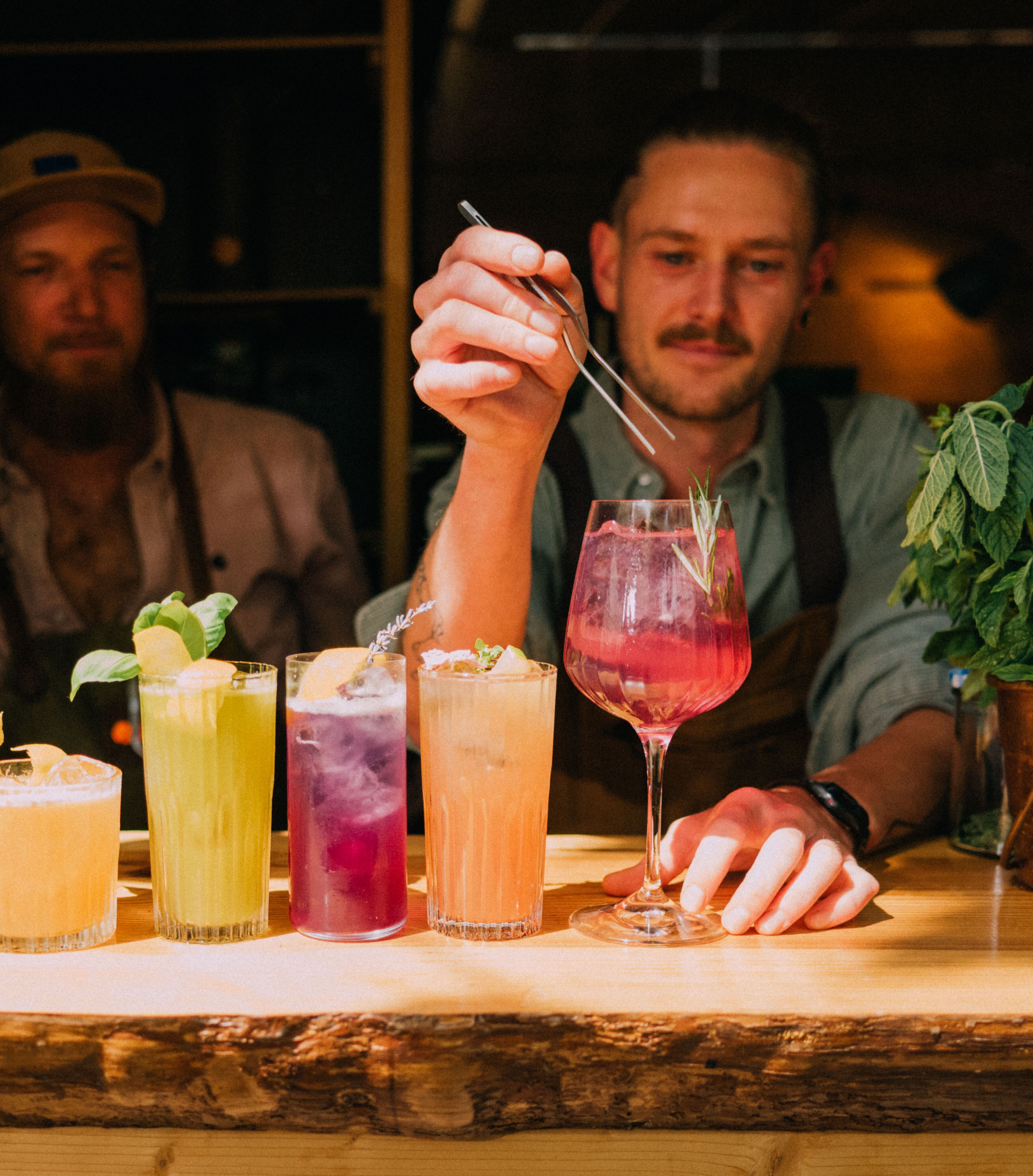 Zwei Barkeeper bereiten an einer Bar fünf farbenfrohe Cocktails zu, umgeben von frischen Kräutern und stilvoller Dekoration.