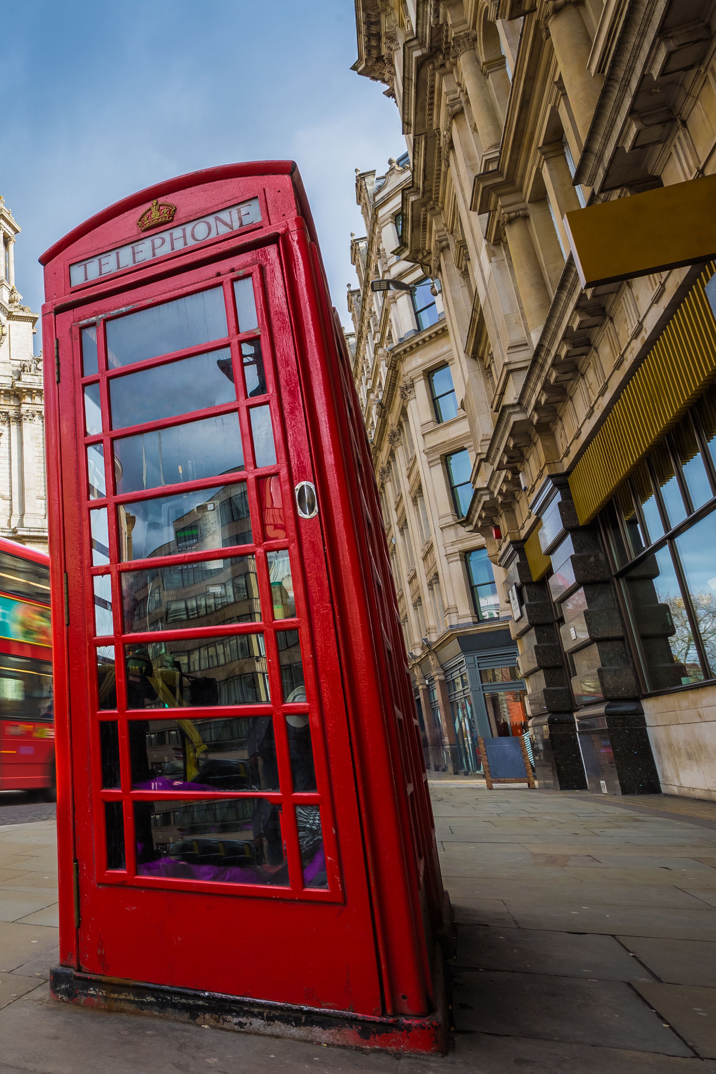 Rote britische Telefonzelle in Nahaufnahme, im Hintergrund ein roter Doppeldeckerbus und die historische Fassade der St.-Pauls-Kathedrale in London unter bewölktem Himmel.