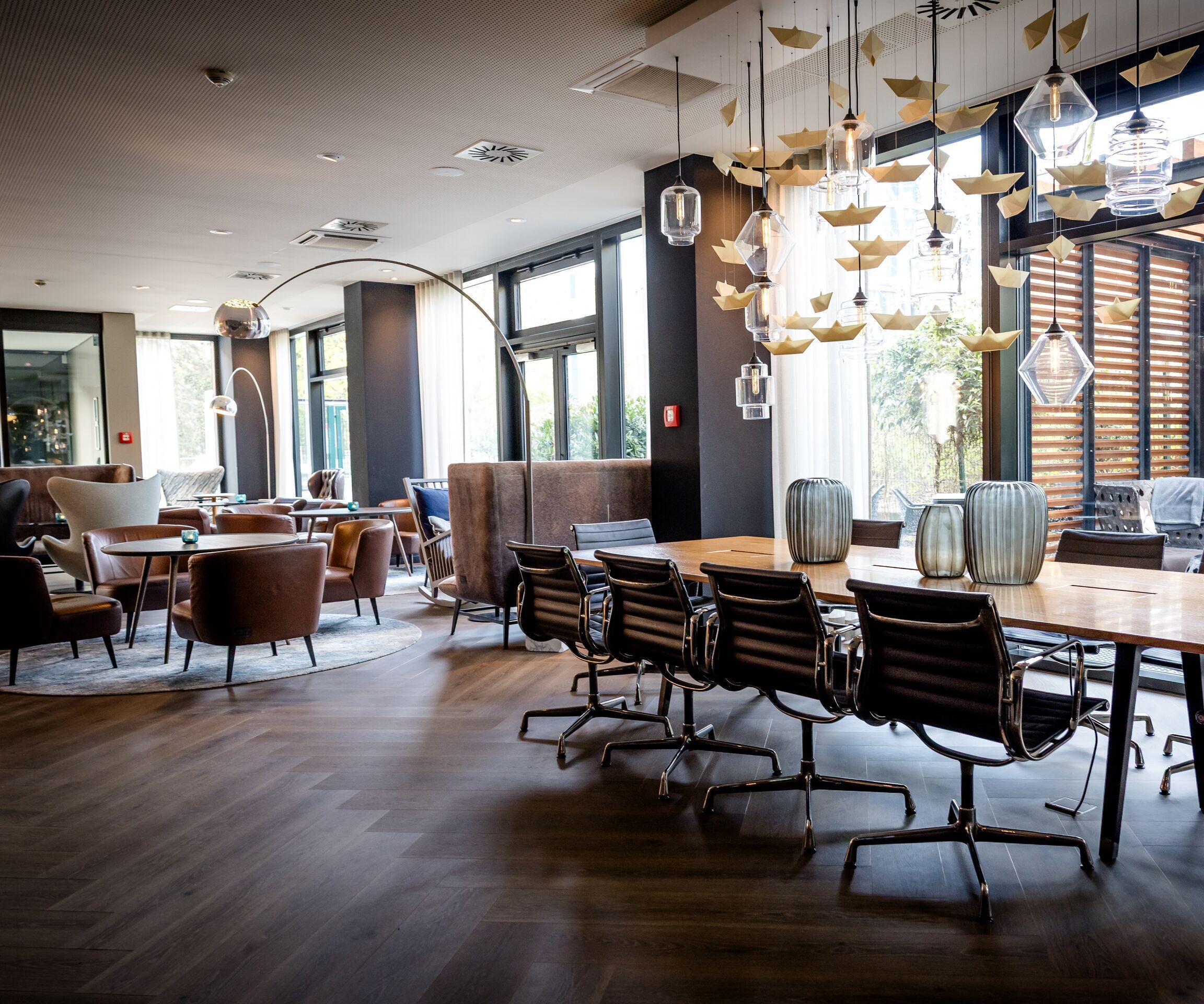 Designhotel Hamburg-Alster Motel One Lounge mit großem Holztisch, Stühlen und dekorativer Hängeleuchte. Große Fenster bieten Blick auf eine Terrasse mit Lounge-Möbeln.