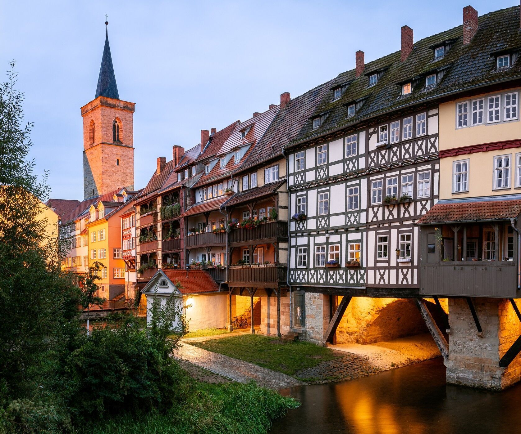 Abendliche Ansicht historischer Fachwerkhäuser, die auf einer Brücke über einen ruhigen Fluss stehen, mit warmem Licht und einem Kirchturm im Hintergrund, vermittelt eine einladende, malerische Atmosphäre einer charmanten Altstadt.