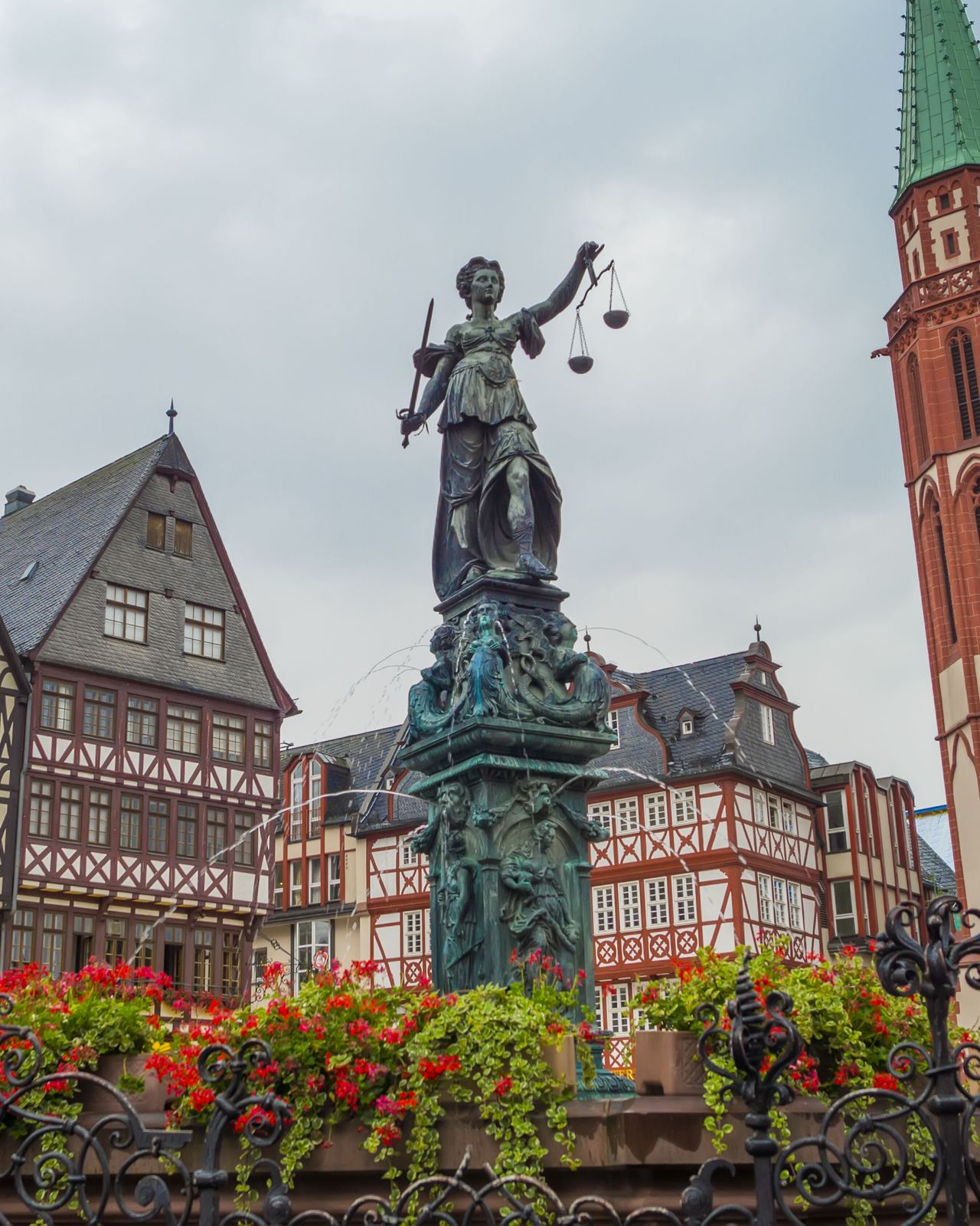 Marktplatz mit Fachwerkhäusern und der Statue der Justitia im Vordergrund, daneben ein hoher Kirchturm mit grünem Dach, an einem klaren Tag.