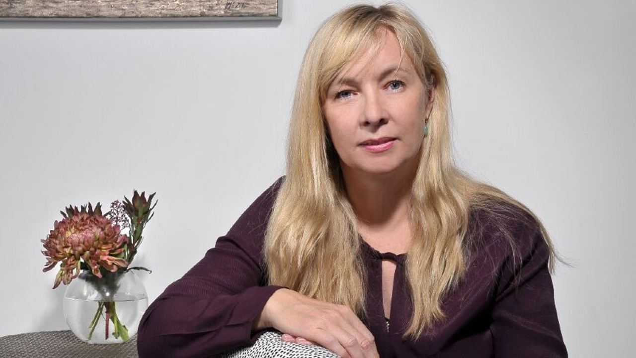 Frau mit blonden Haaren sitzt entspannt auf einem gepolsterten Stuhl, neben ihr ein kleiner Tisch mit einer Vase und Blume.