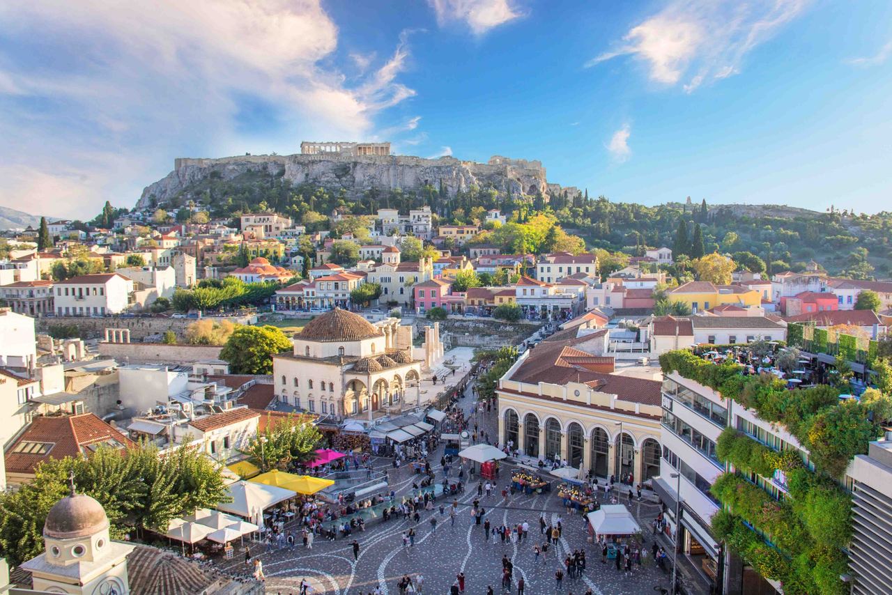 Panoramablick auf die historische Altstadt von Athen mit belebtem Marktplatz und der Akropolis im Hintergrund unter blauem Himmel.
