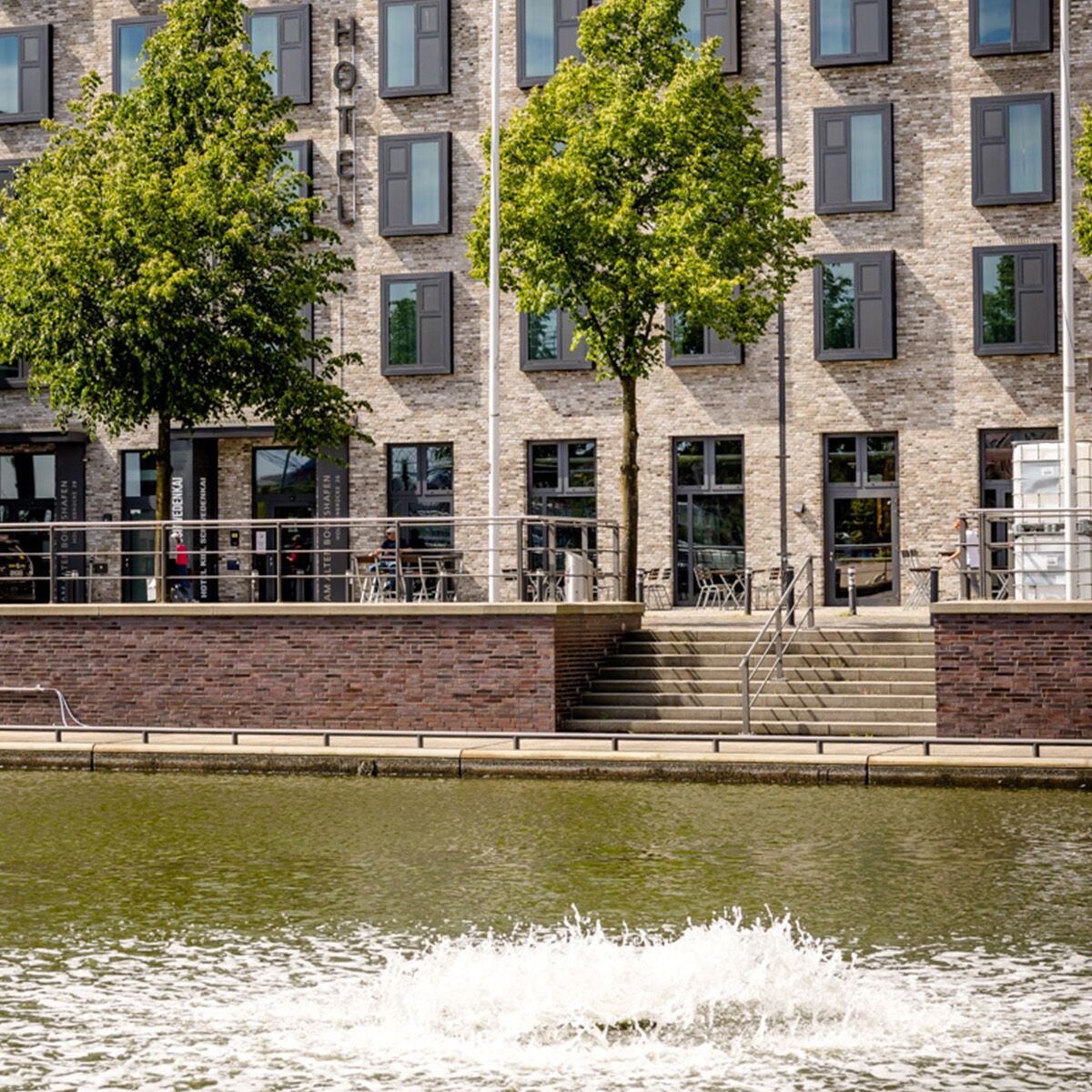 Fassade eines modernen Hotels mit vielen Fenstern, davor ein Wasserbecken mit Fontäne und drei grüne Bäume entlang des Ufers, die eine ruhige und einladende Atmosphäre schaffen.